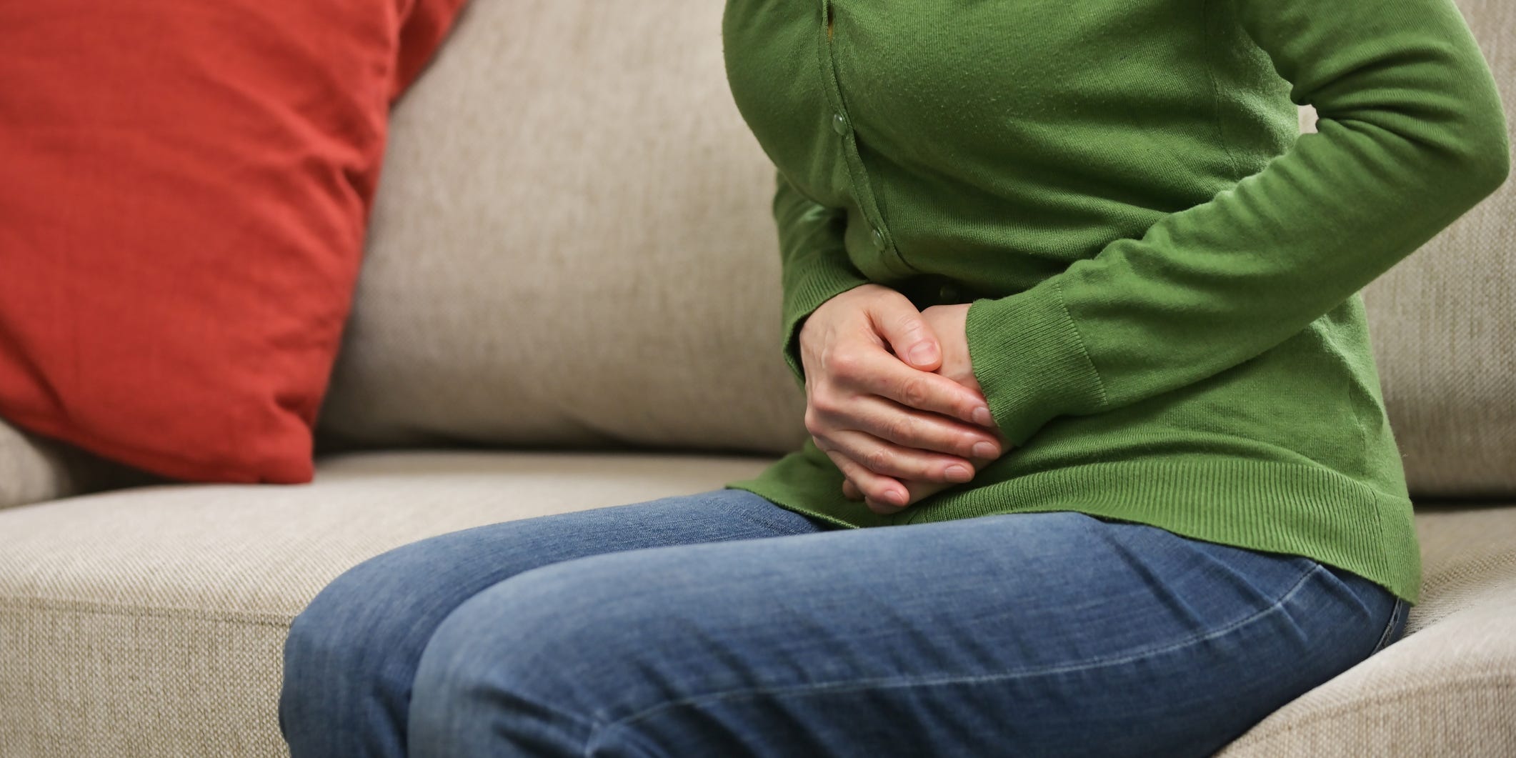 A woman sitting on a couch in a green shirt and jeans clutches her stomach in pain.