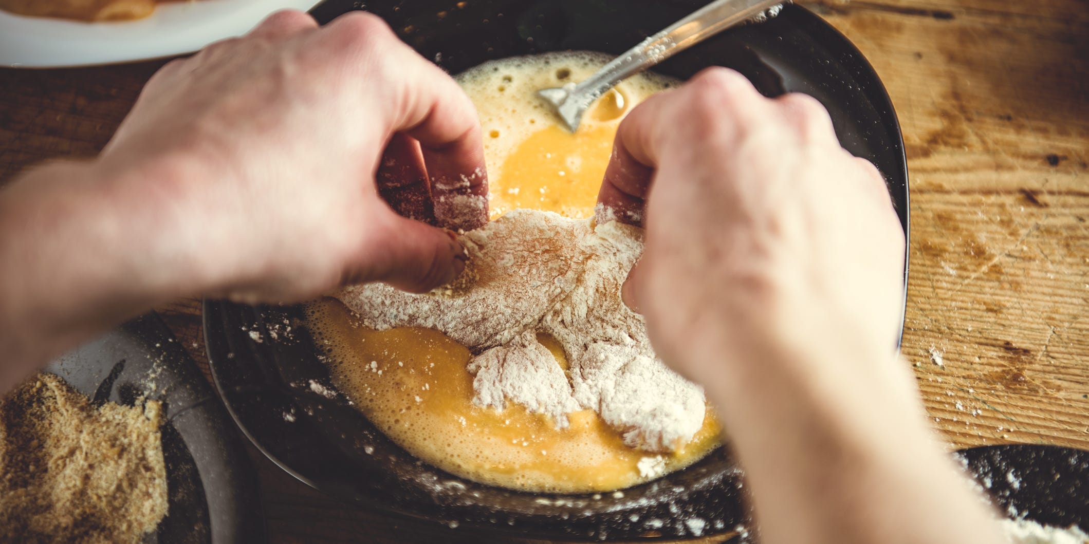 A pair of hands dipping flour-coated chicken into egg wash
