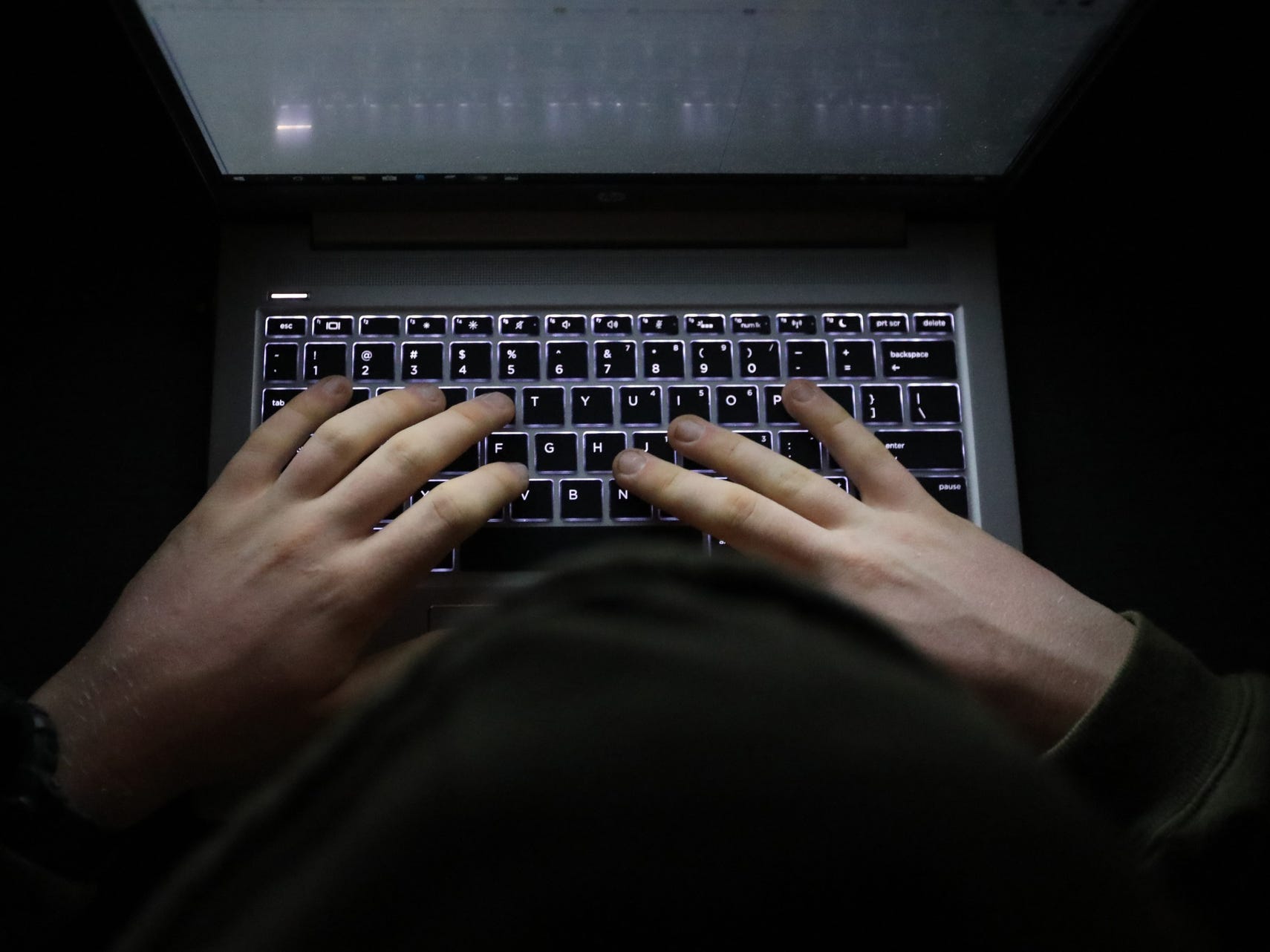 A simple, very dark night time image of hands on an illuminated keyboard typing. Shady person wearing a hood at a computer or laptop in the dark.
