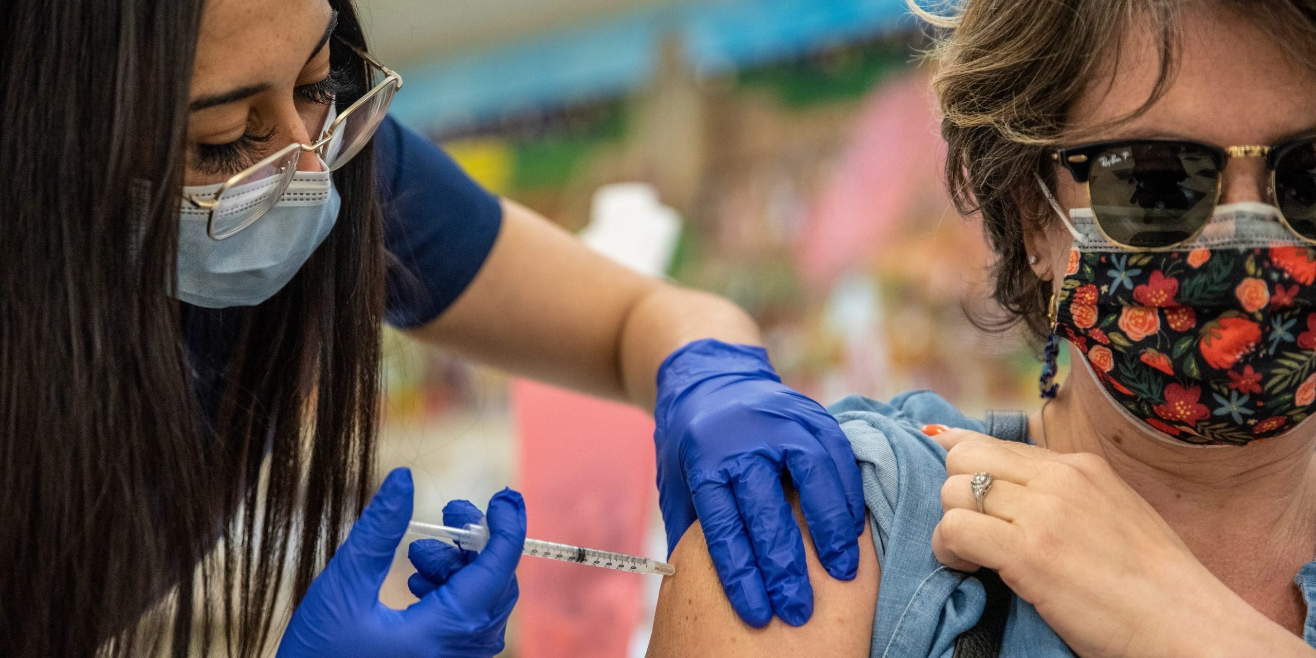 Women with mask and glasses administers COVID-19 vaccine to middle-aged women with sunglasses and patterned mask