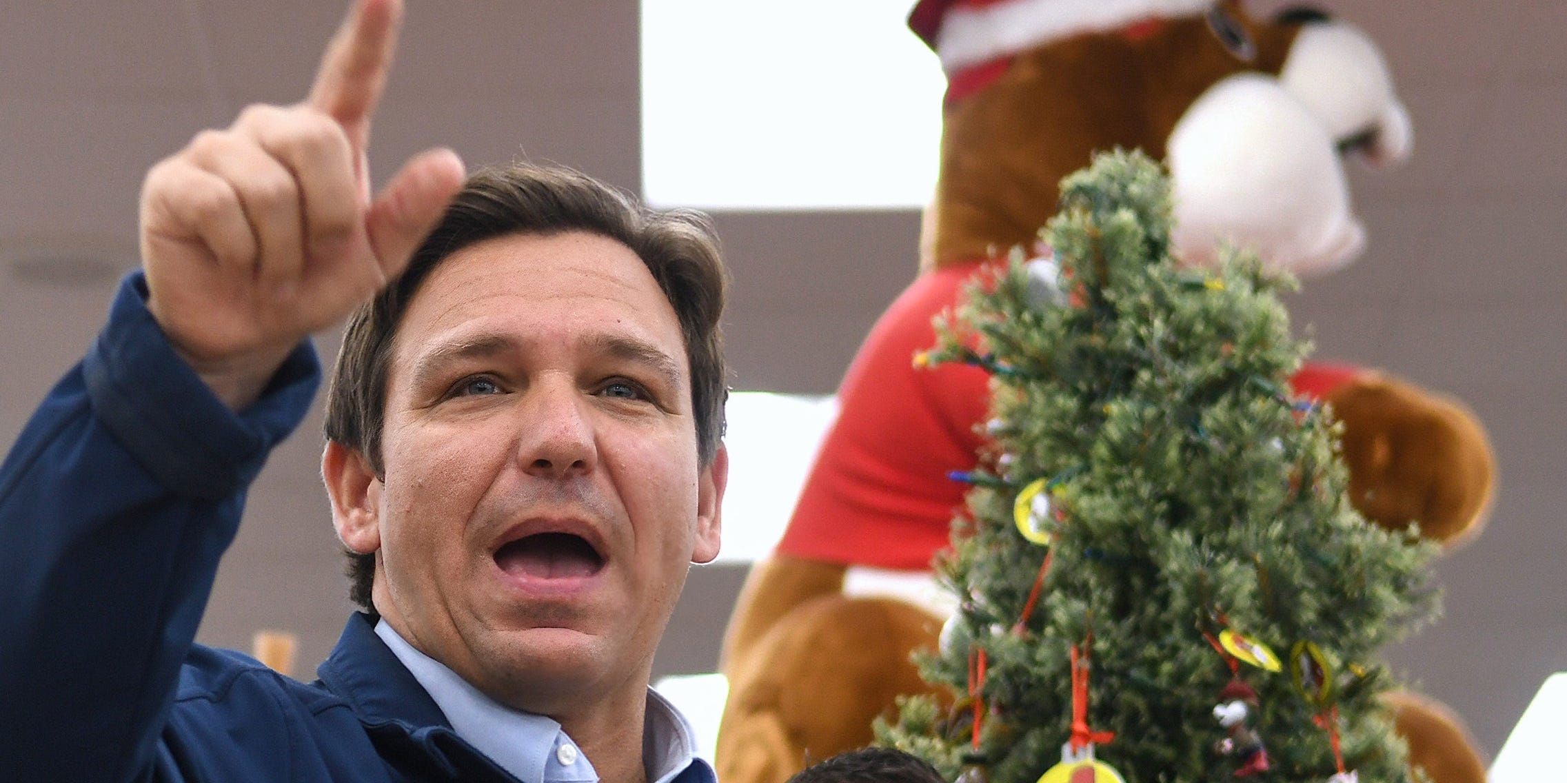 Ron DeSantis points to the ceiling in front of a Christmas tree with a bear on it