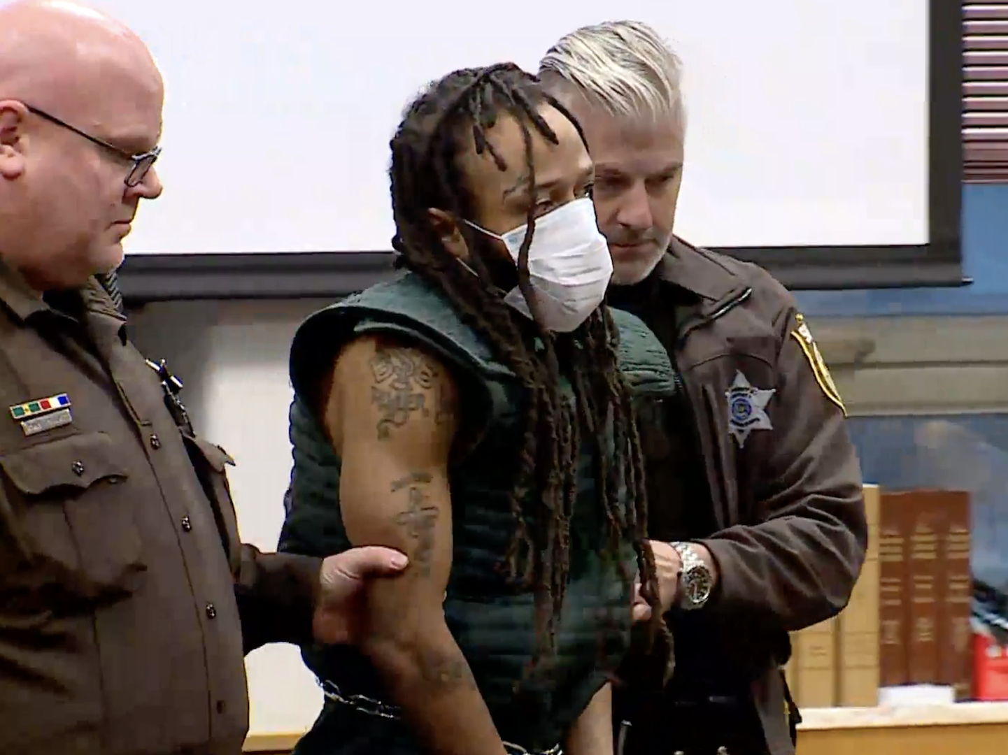 A still from courtroom video showing Darrell Brooks, masked and in handcuffs, escorted by two officers into his hearing on November 23, 2021