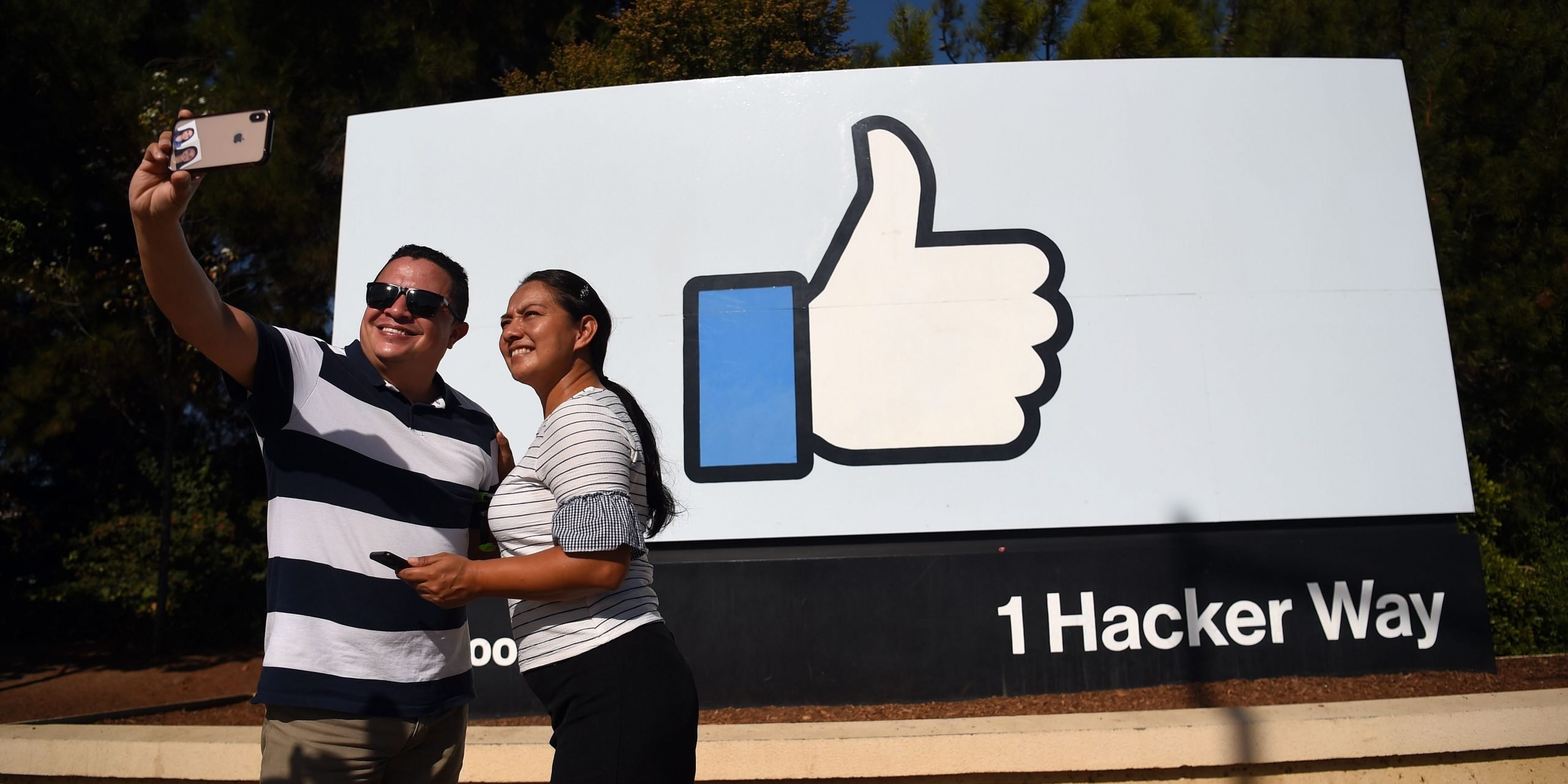 Two people taking a selfie in front of the Facebook "thumbs up" sign at the company's heaequarters