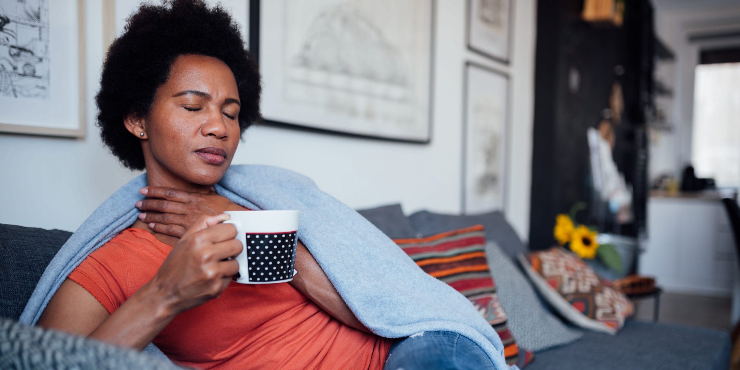 A woman sits on a couch and holds a mug of tea as she massages her throat.