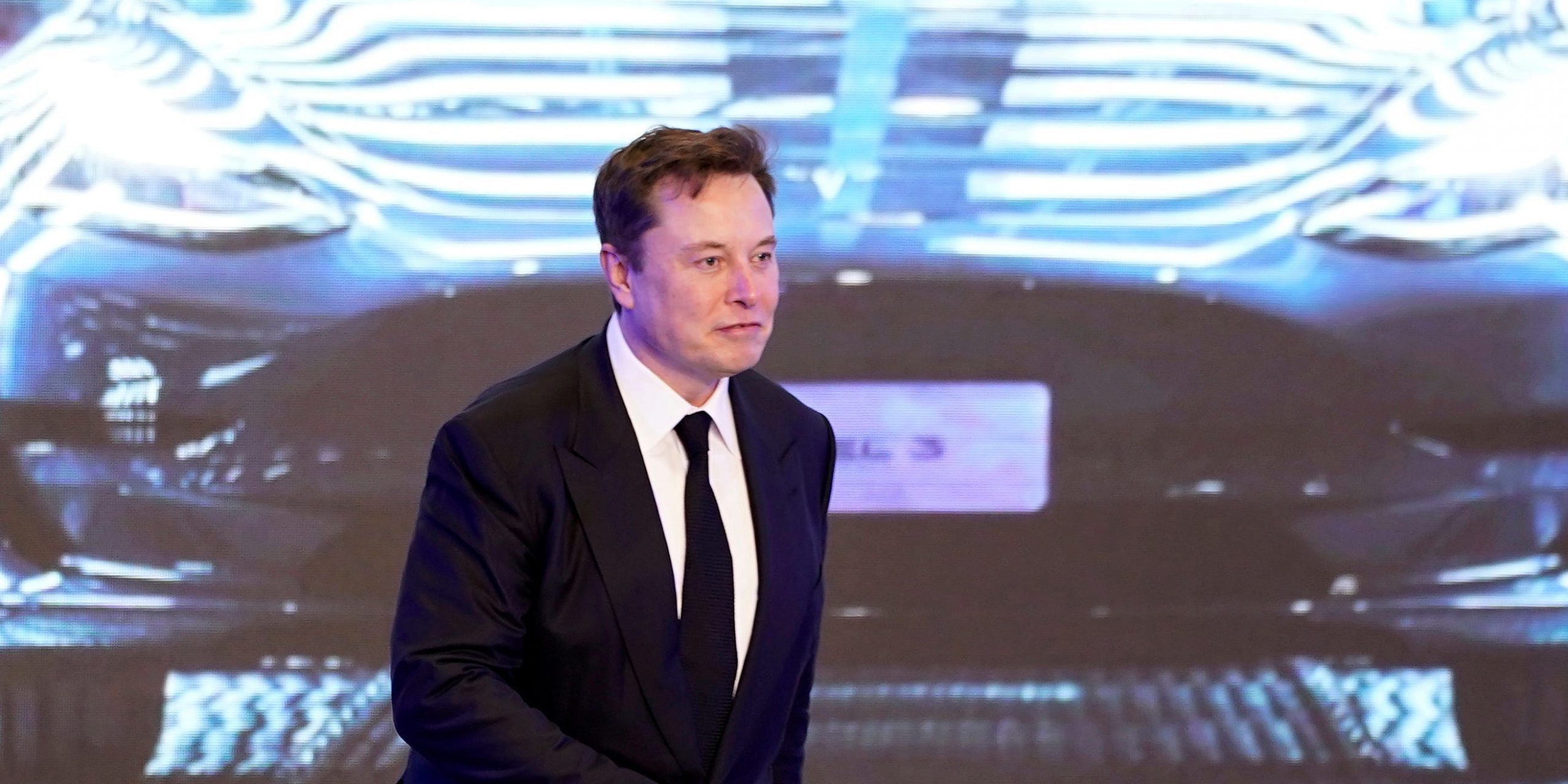 Tesla CEO Elon Musk in a black suit walks on stage in front of an image of a Model Y vehicle