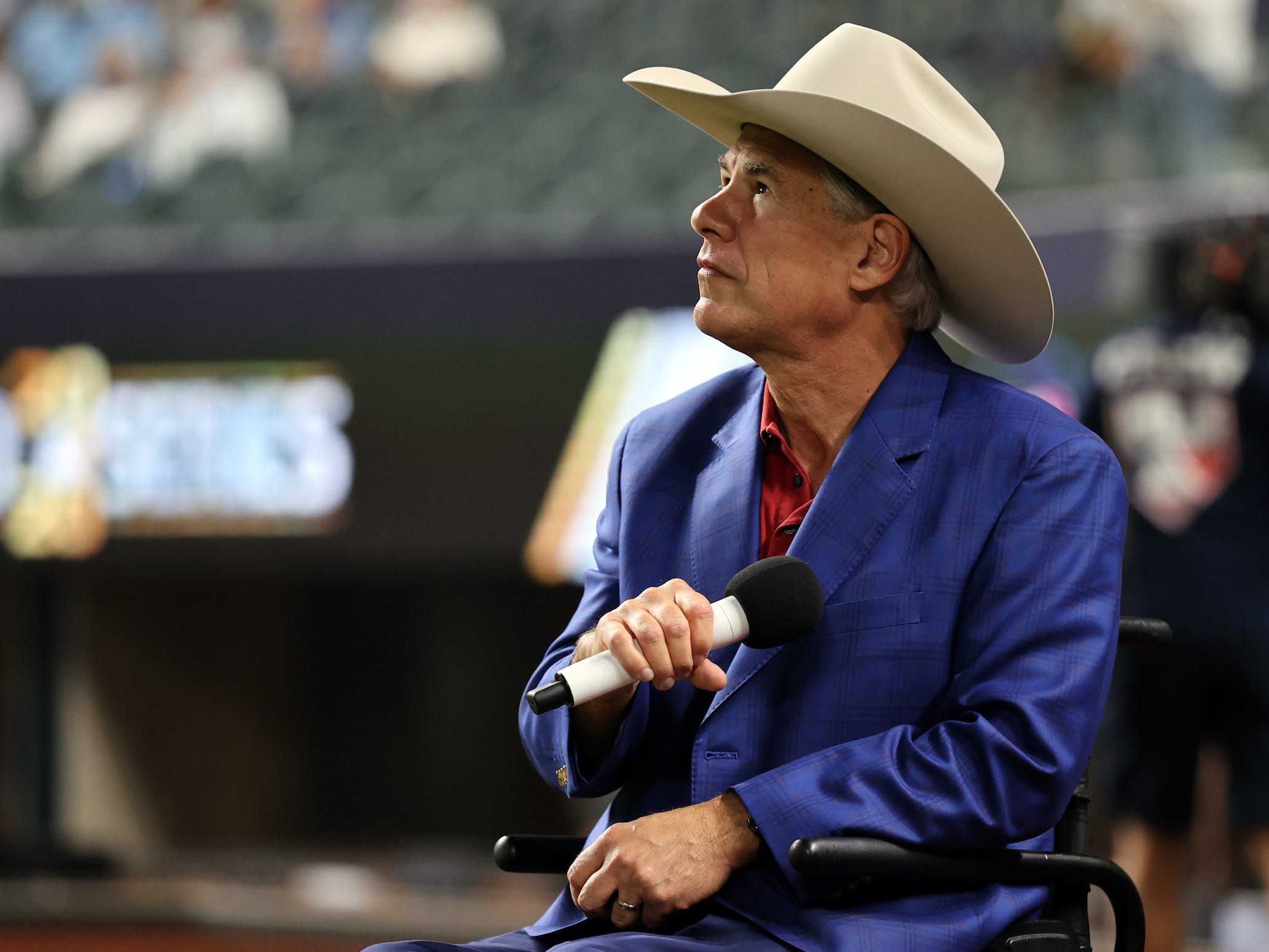 Wearing a tan cowboy hat, Texas Governor Greg Abbott lowers a microphone toward his chest while listening to the national anthem at a World Series baseball game held in Arlington, Texas.