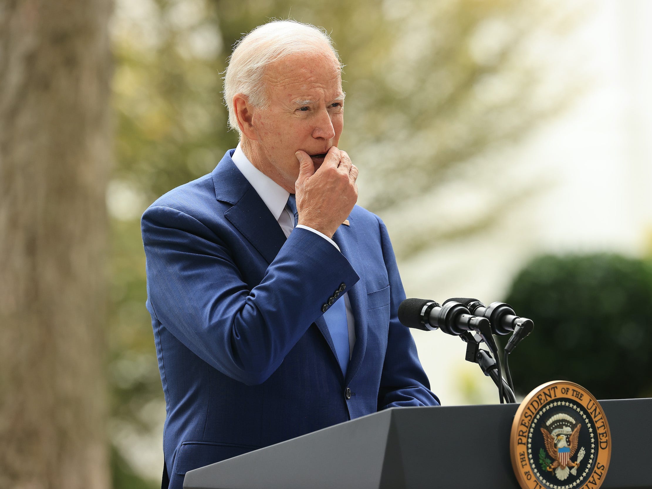 President Joe Biden stands outside behind a podium with the presidential seal, grabbing his chin and slightly covering his mouth in a pensive look.