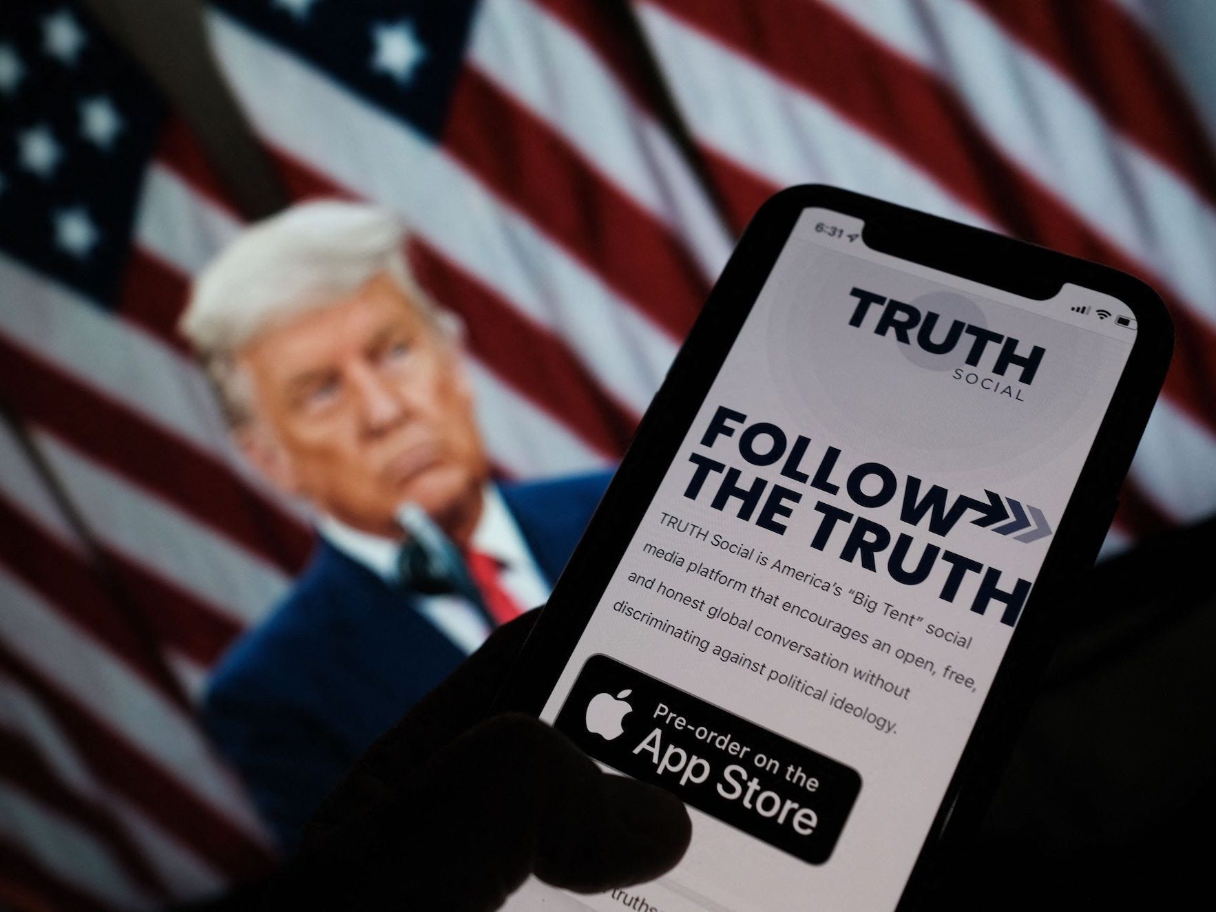 A photo illustration showing a person checking the app store on a smartphone for "Truth Social", with a photo of former President Donald Trump in the background.