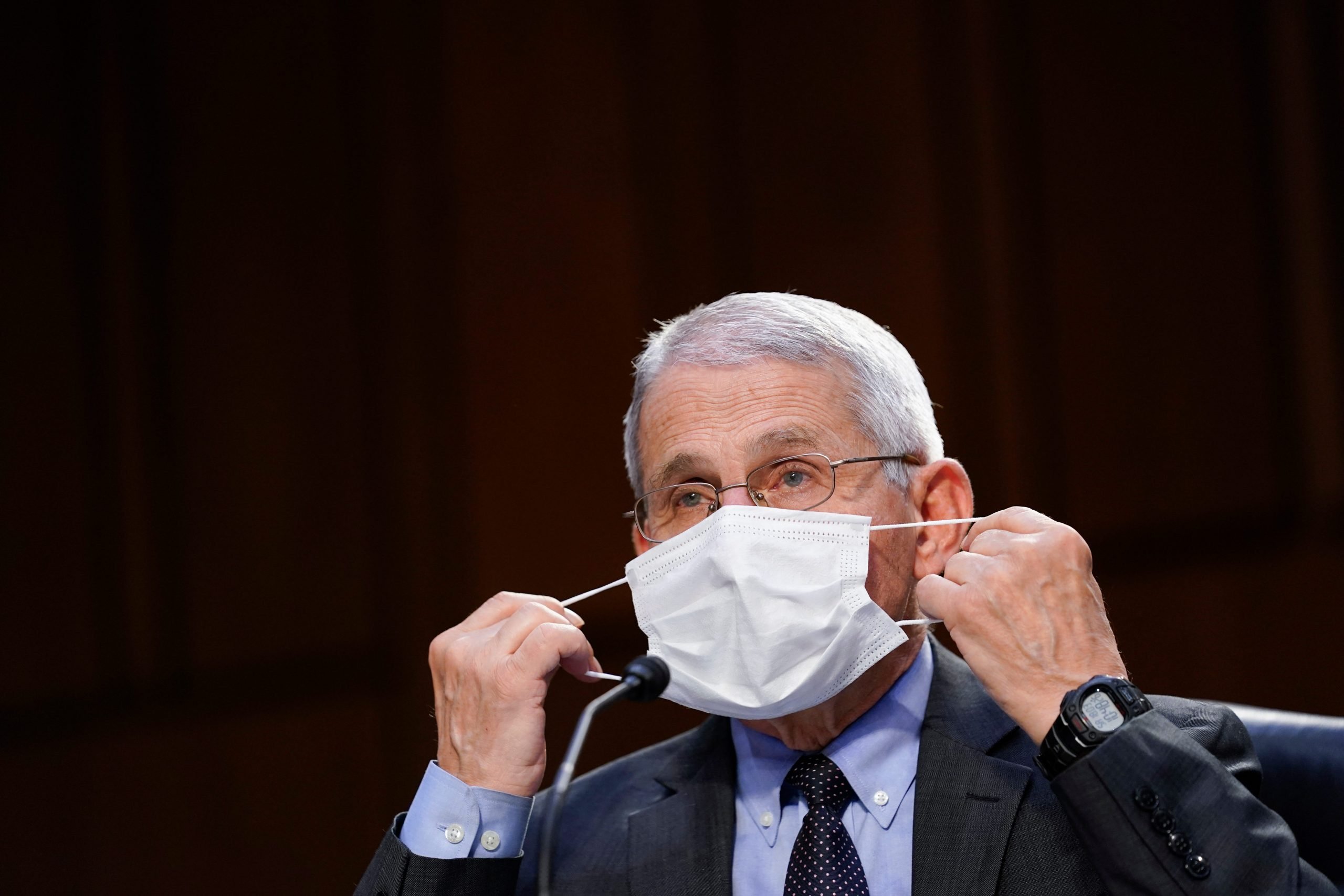 Dr. Anthony Fauci puts on white mask, dressed in suit. Black background.