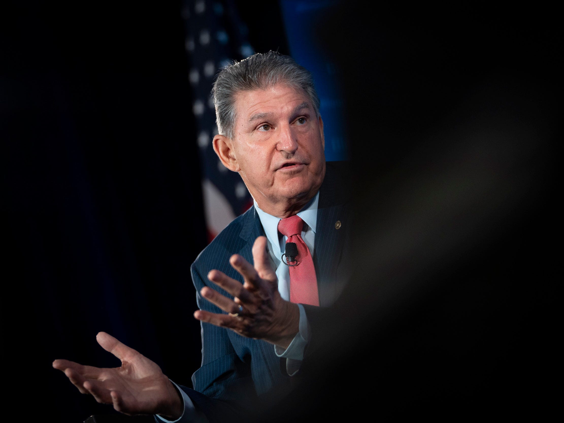 Senator Joe Manchin gestures in front of an American flag with his palms up, the right half of the photo is blurred out in black.