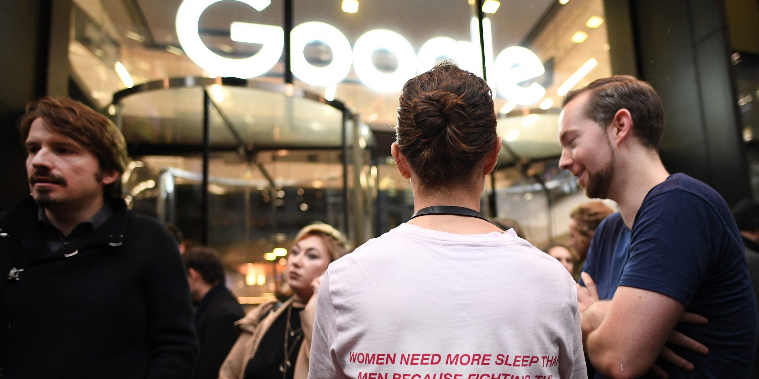 A woman in front of a Google sign on a building, stands with her back facing the camera. On the back of her T-shirt it says "Women need more sleep than men because fighting the patriarchy is exhausting."