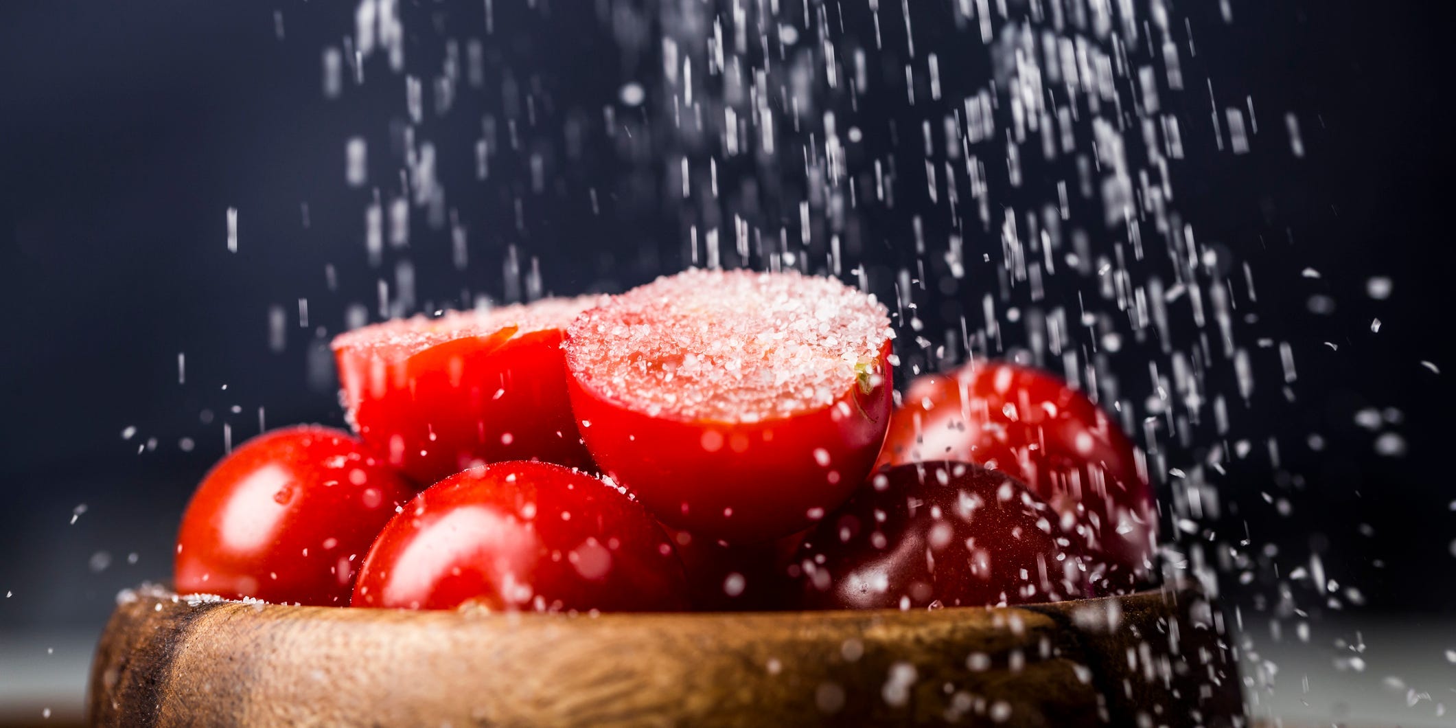 Salt raining down on a wooden bowl of halved cherry tomatoes