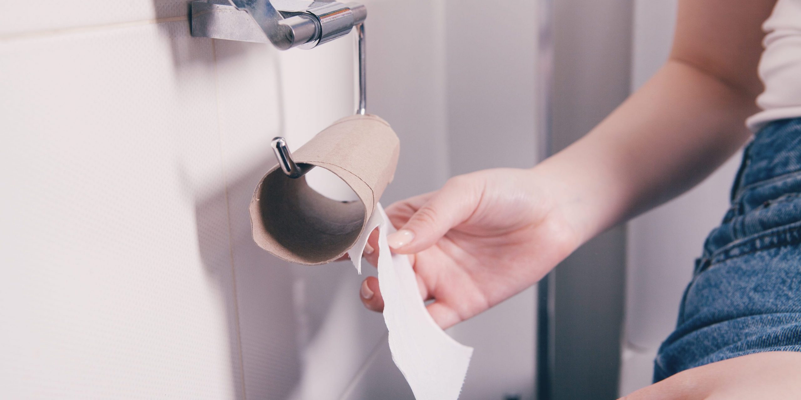 A woman sitting on a toilet holds the last sheet of toilet paper on a roll.