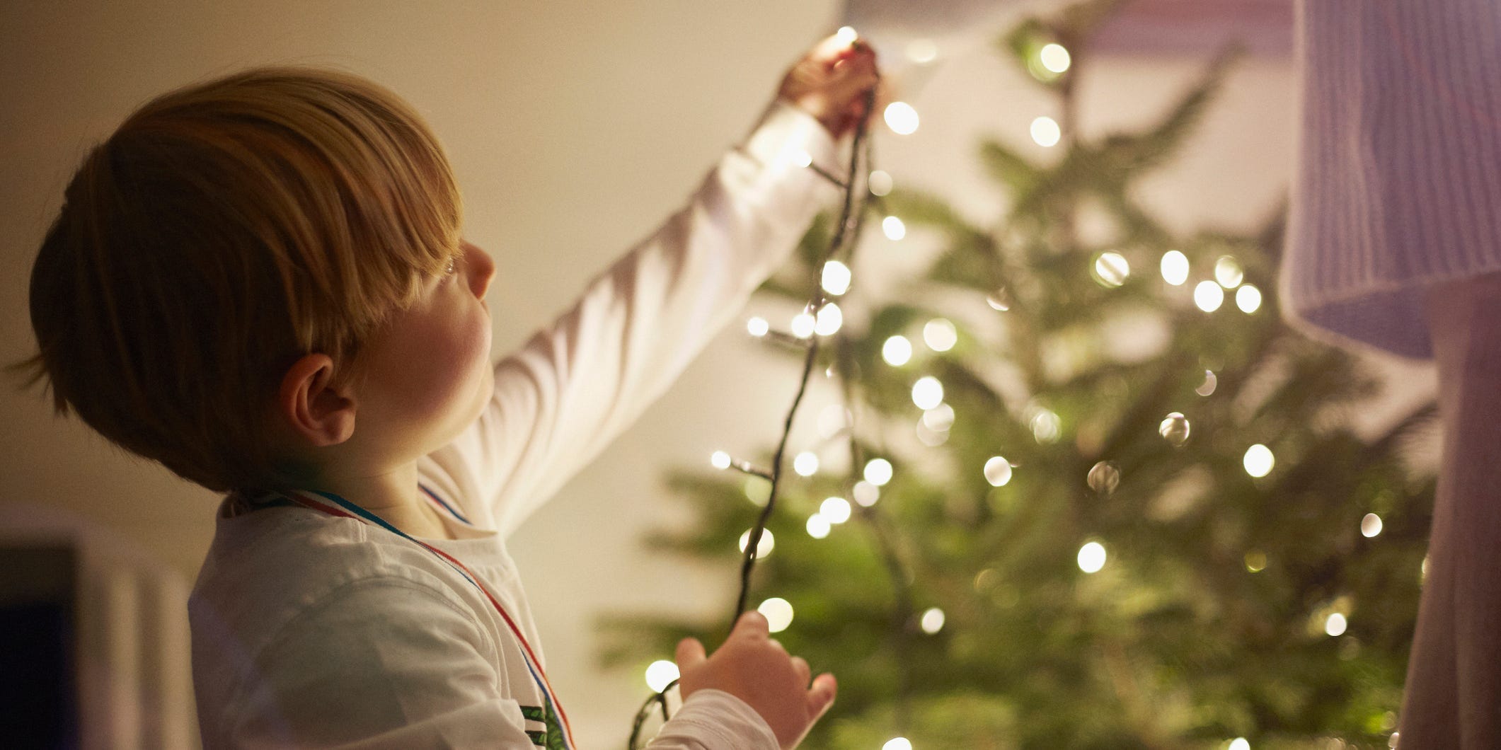 A young child adding white string lights to a Christmas tree