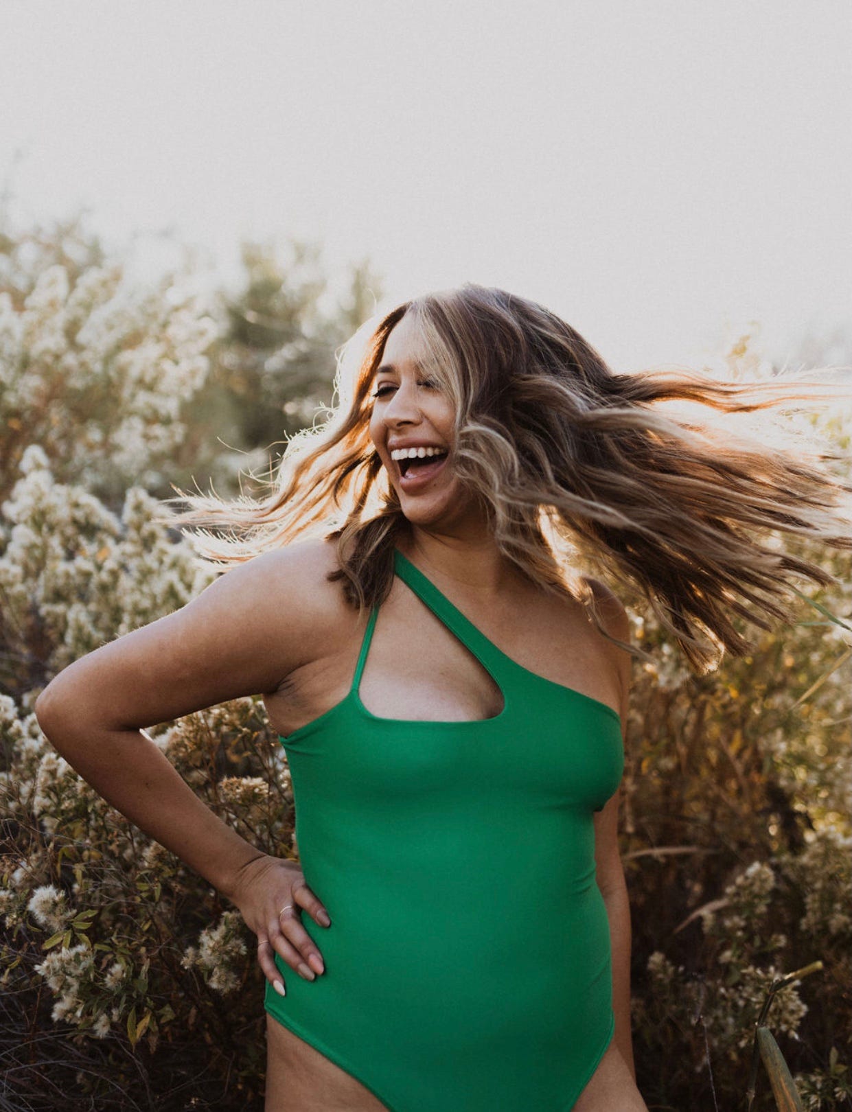 Woman wearing green Arrow + Phoenix swimsuit laughs outdoors with hand on hip