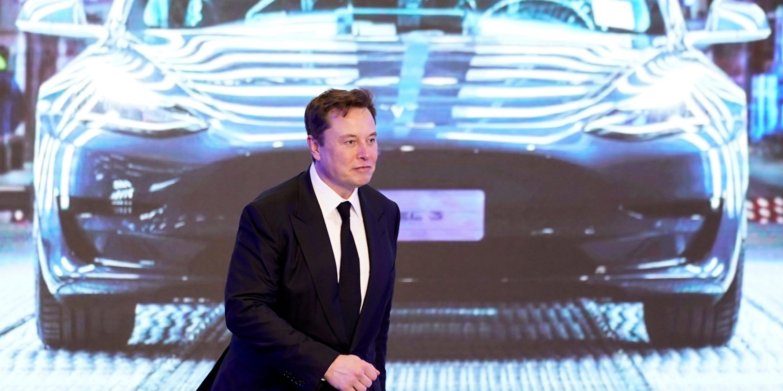 Tesla CEO Elon Musk in a black suit walks on stage in front of an image of a Model Y vehicle
