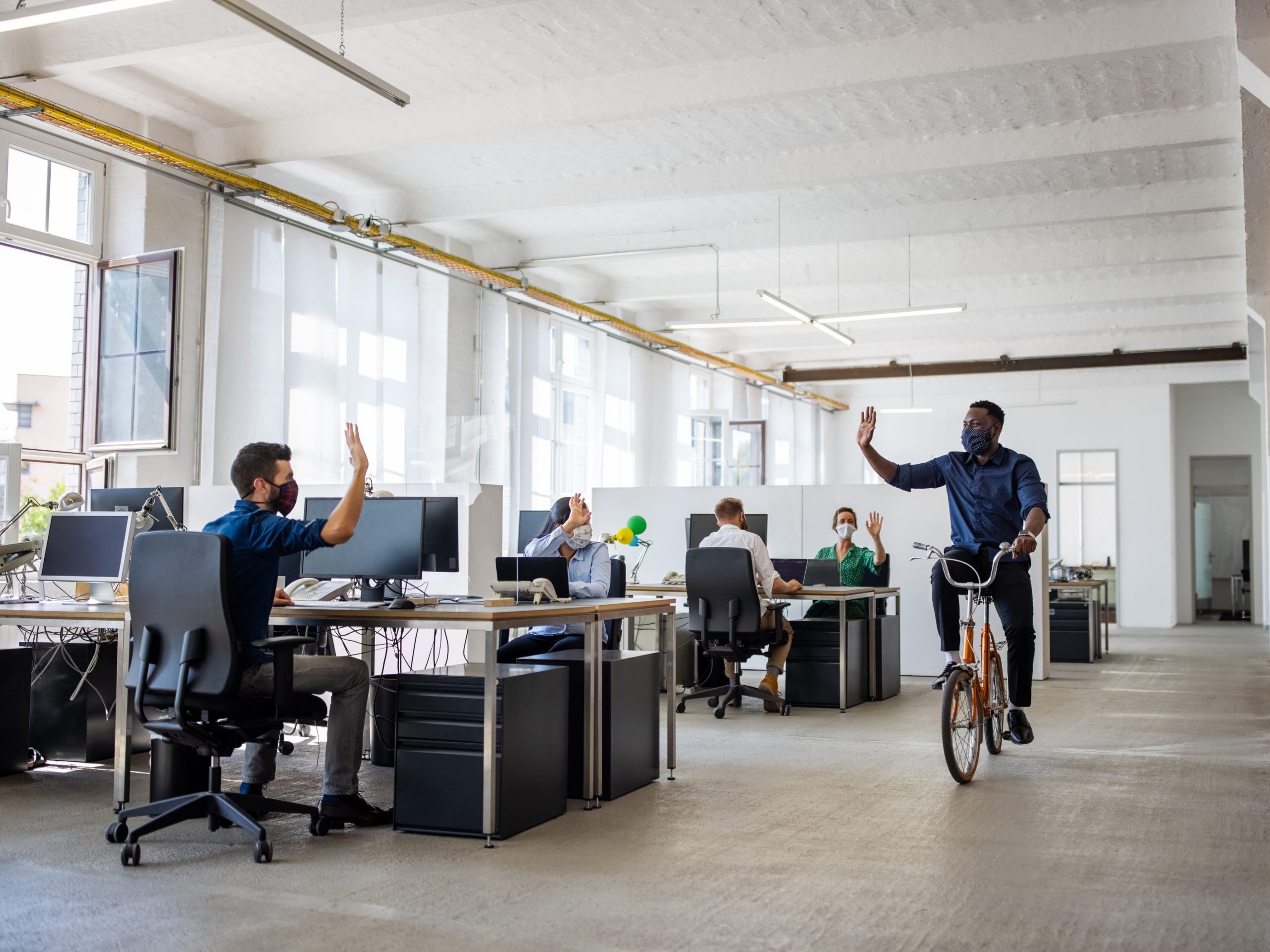 A man ride a bicycle through an office while waving to his coworker.