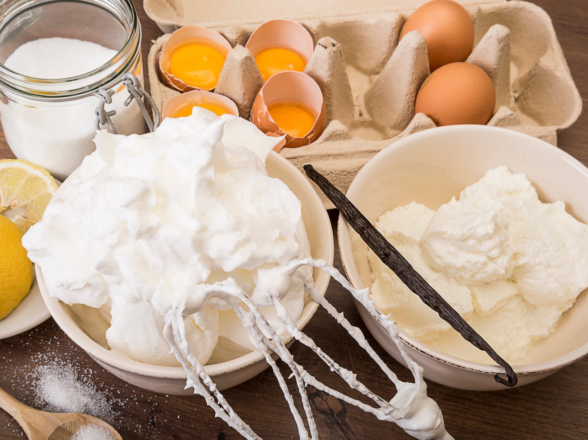 Bowl of beaten egg white and other baking ingredients of meringues on wooden table.