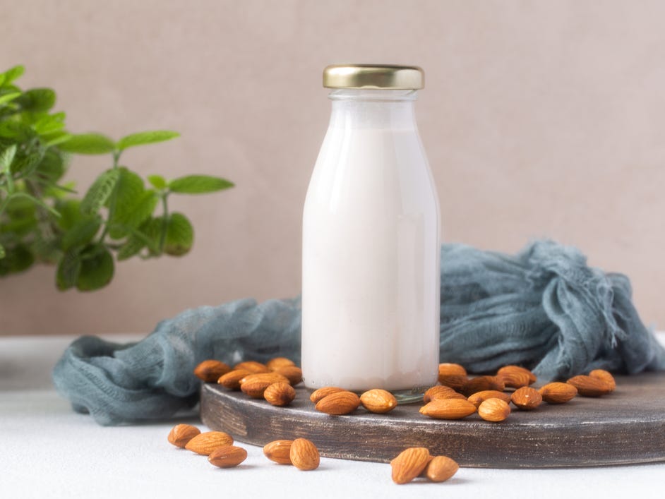 Almond milk in glass bottle with almond nuts on wooden cutting board and houseplant on background.