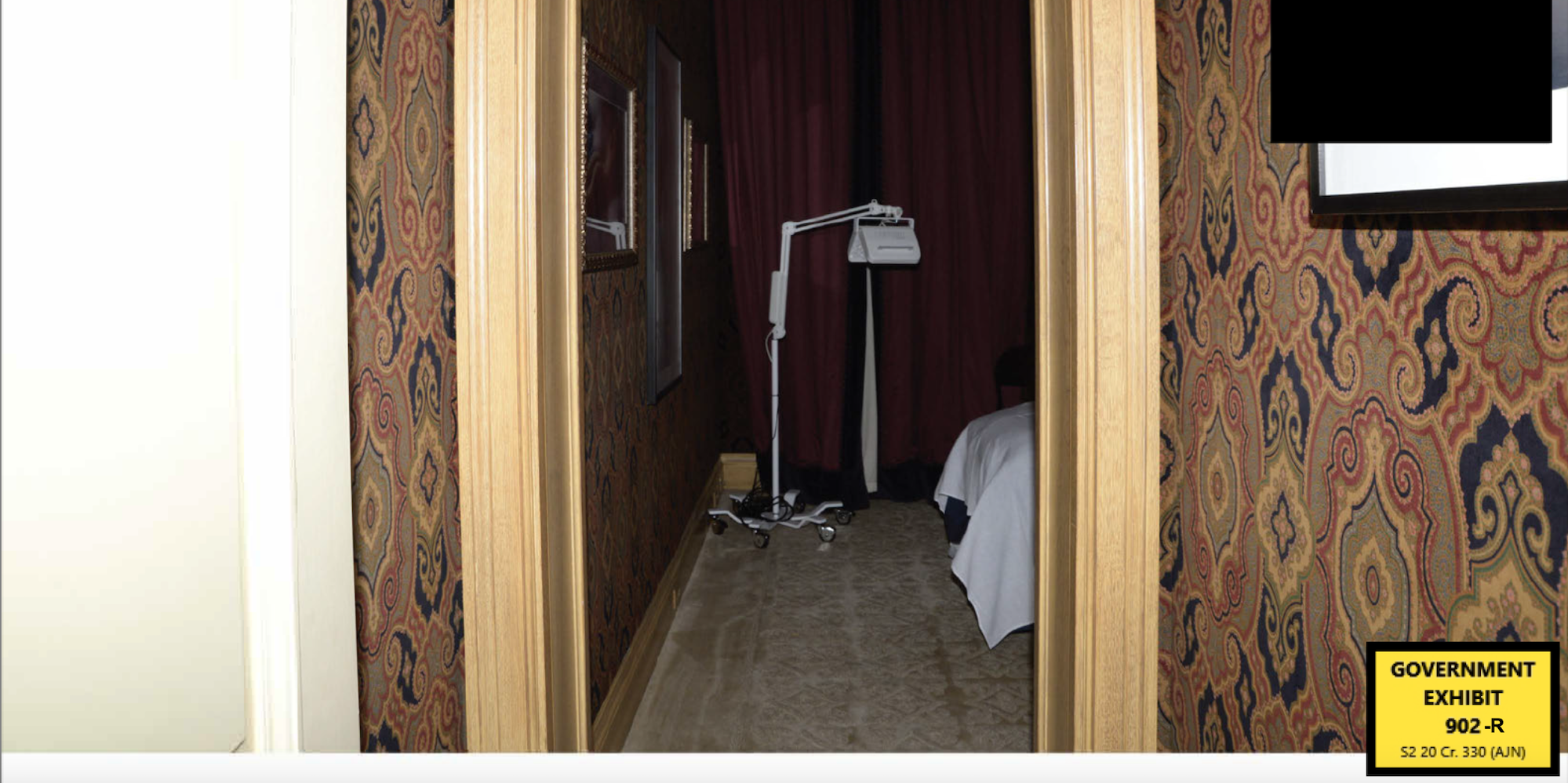 A photo, labeled as a government exhibit, showing a doorway leading to a darkened room. The edge of a table, covered in a white cloth, can be seen. A picture in the hallway has been redacted with a black square.