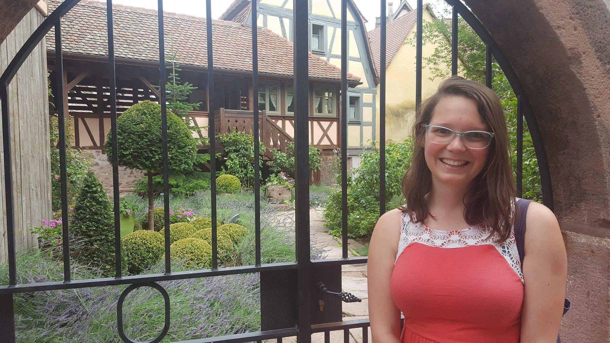 The author, Taryn Williams, standing in front of a gate in an orange shirt. She has short brown hair and blue glasses.