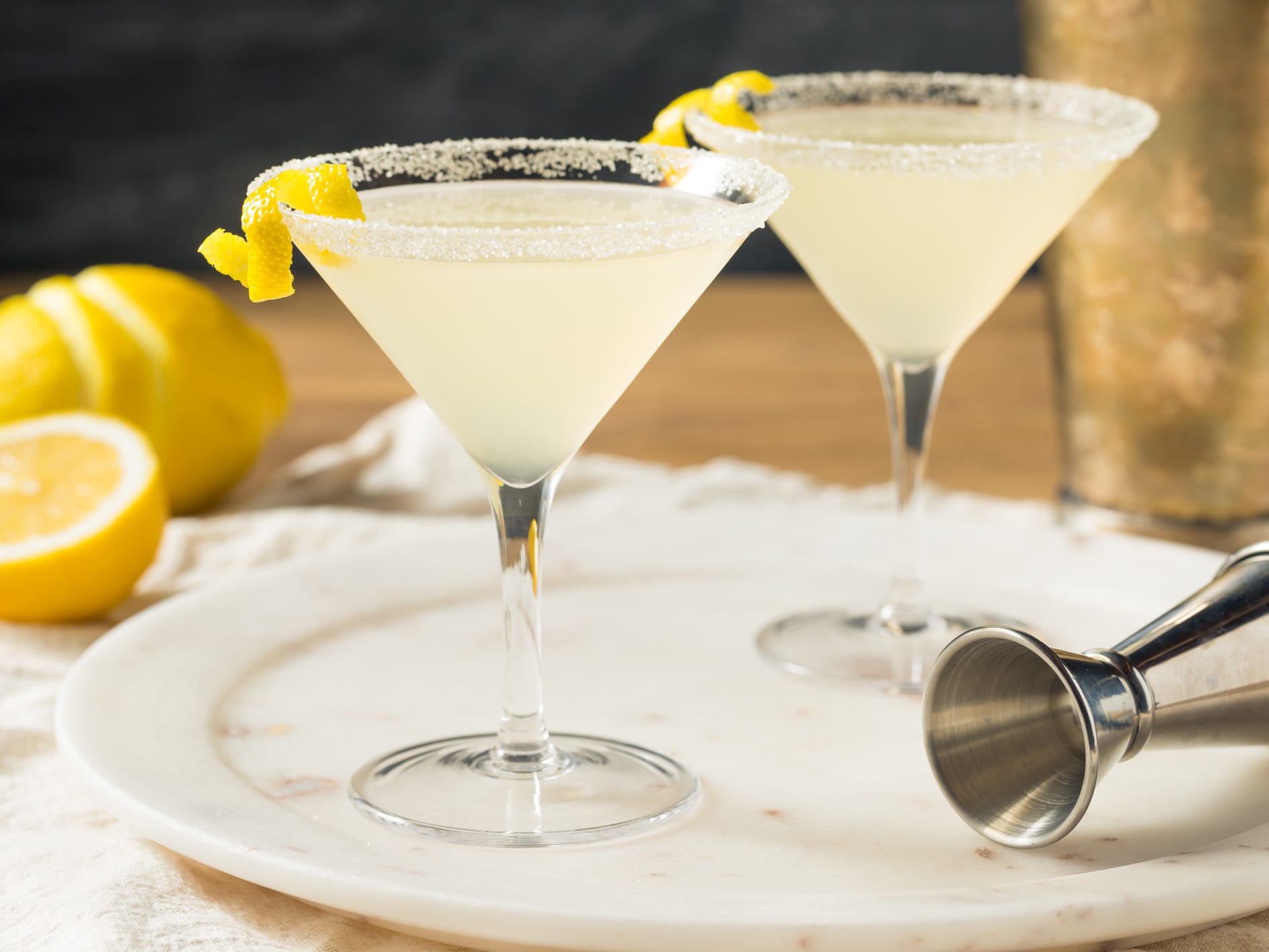 Two lemon drop cocktails in martini glasses surrounded by a zested lemon and a cocktail jigger