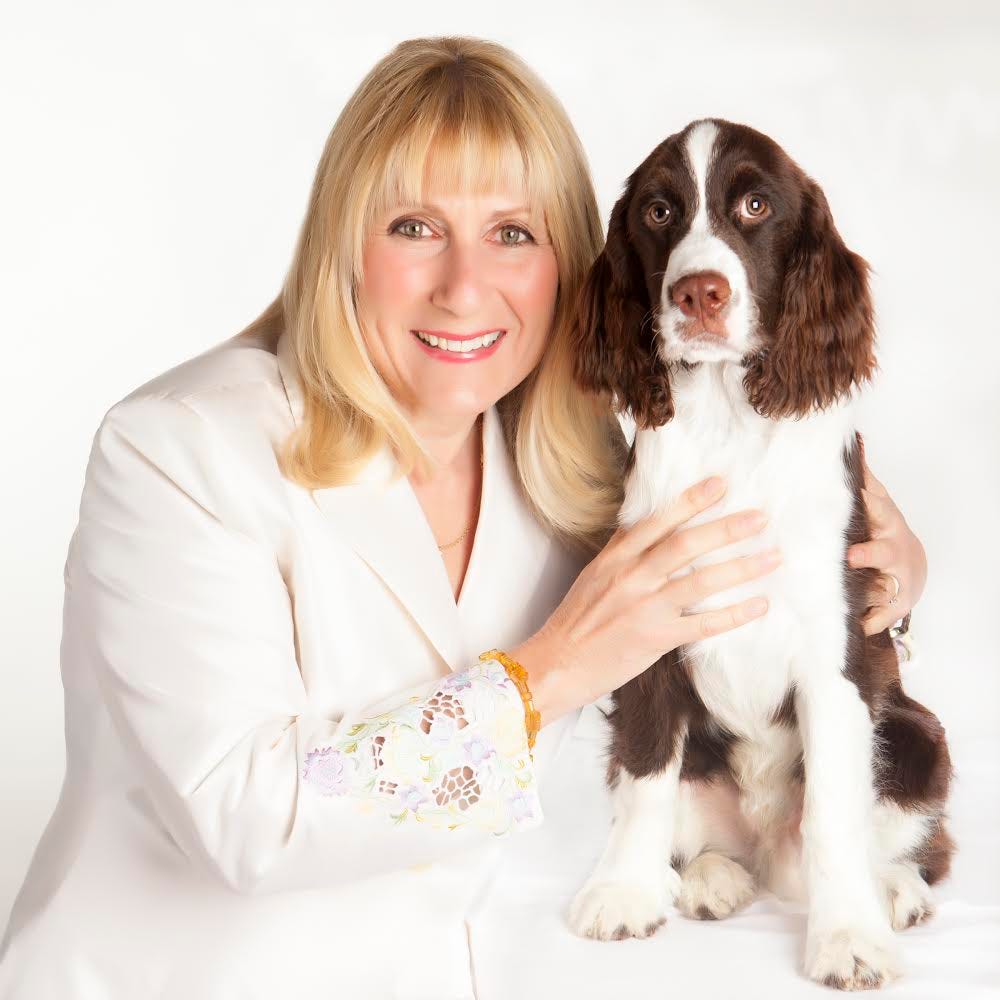 Janet Marlow, who has blonde hair and a white shirt on, smiling next to a brown and white dog.