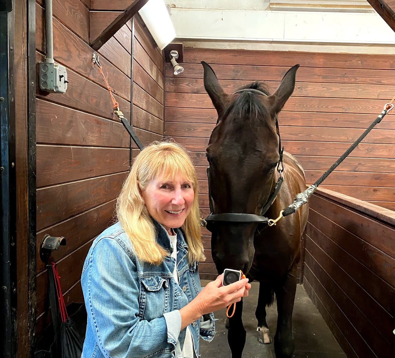 Janet Marlow, who has blonde hair and a denim jacket on, smiling next to a horse in a stable.