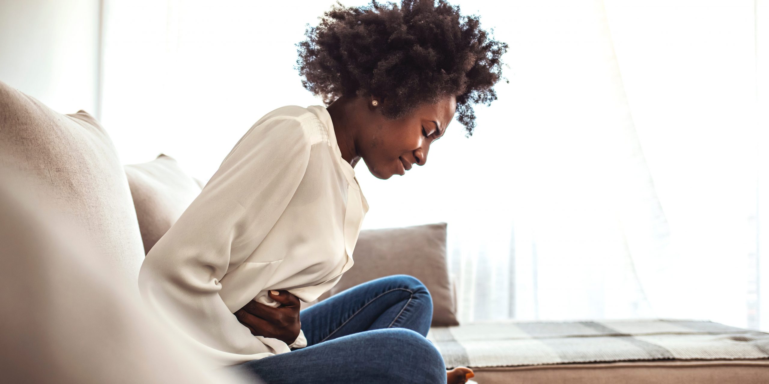 A woman sitting on a couch curls over and holds her hips and stomach in pain.