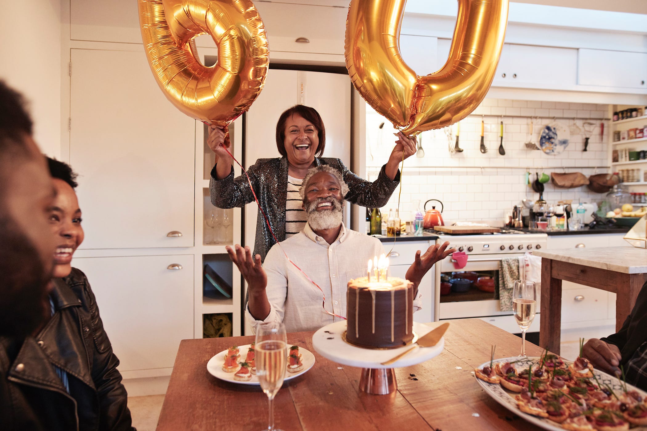woman with '60' baloons standing behind smiling man with cake, celebrating his 60th birthday at a table with friends in the kitchen