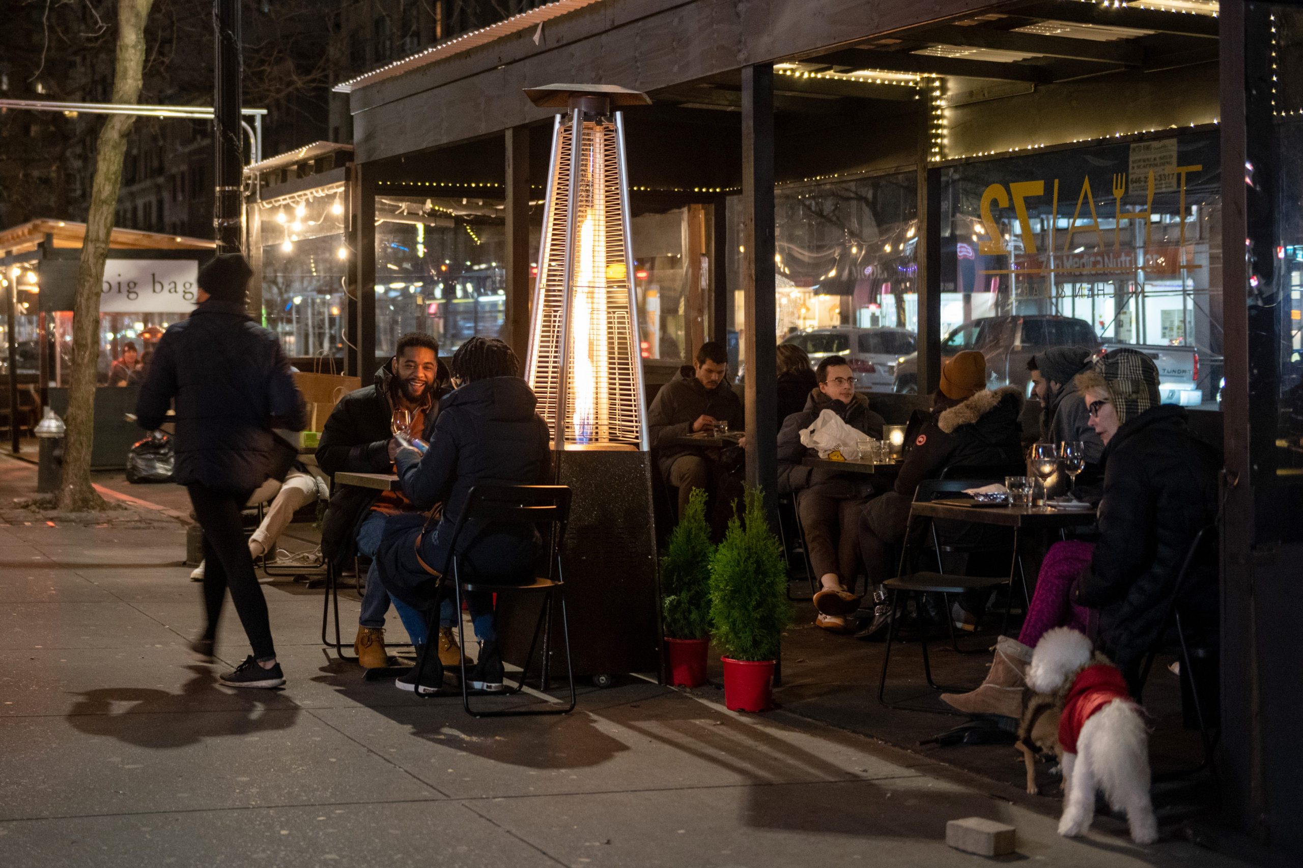 People are seen dining at sidewalk tables outside next to a tall outdoor heater.