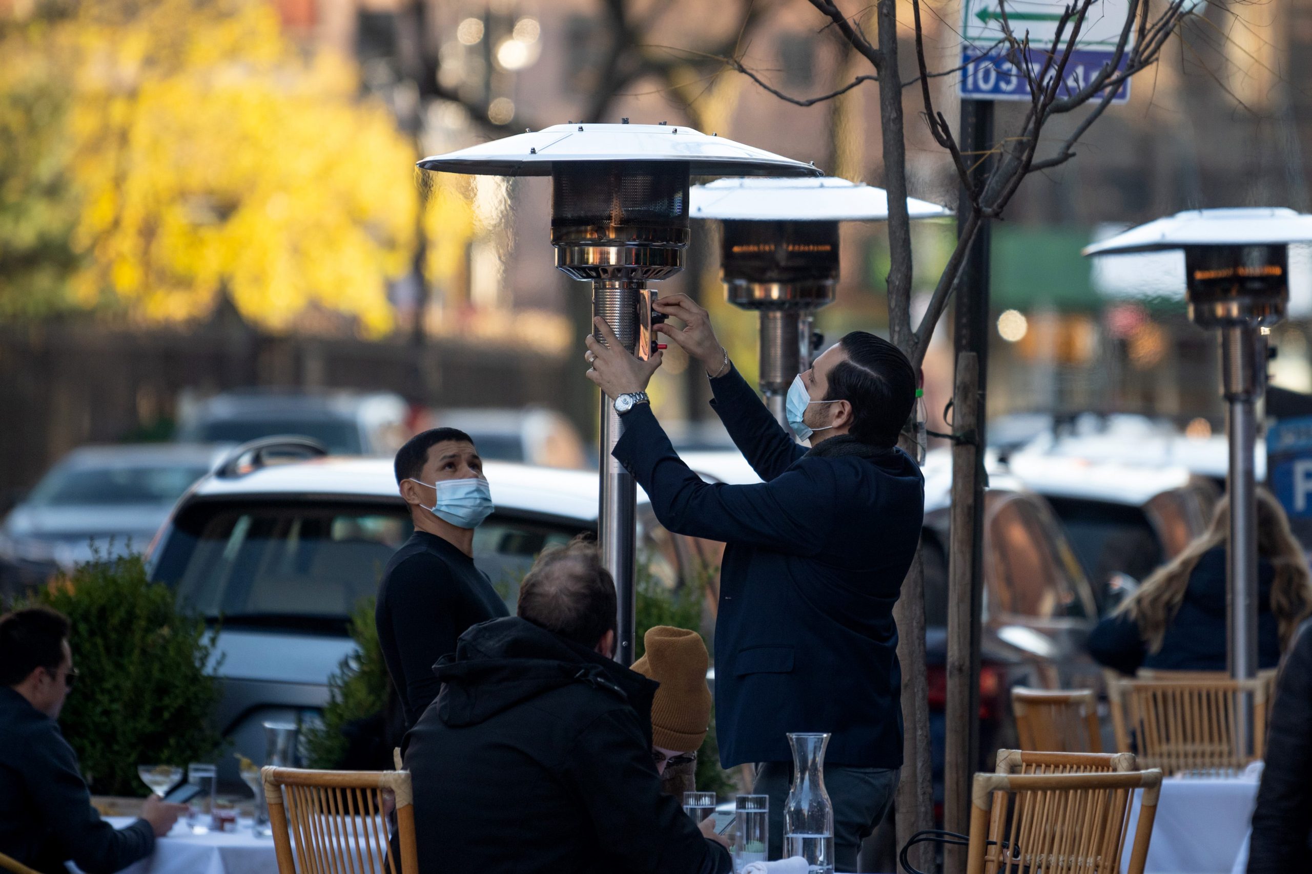 A masked restaurant worker adjusts an outdoor propane heater as two people, seated, look on.