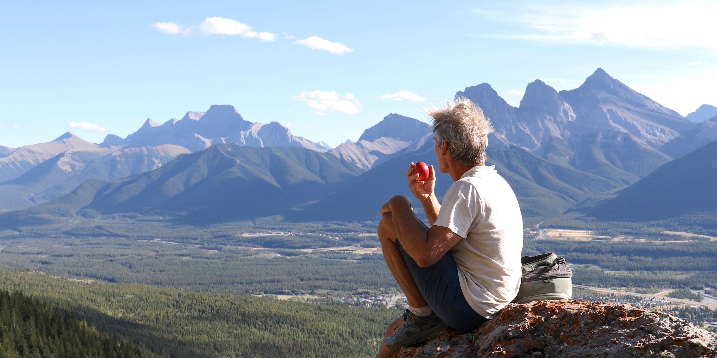 A man sitting, eating an apple mindfully while he looks onto a vast mountainous landscape.