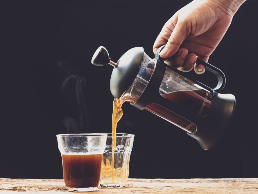 Coffee is poured into a cup from French press coffee maker on an old wood table and black background.