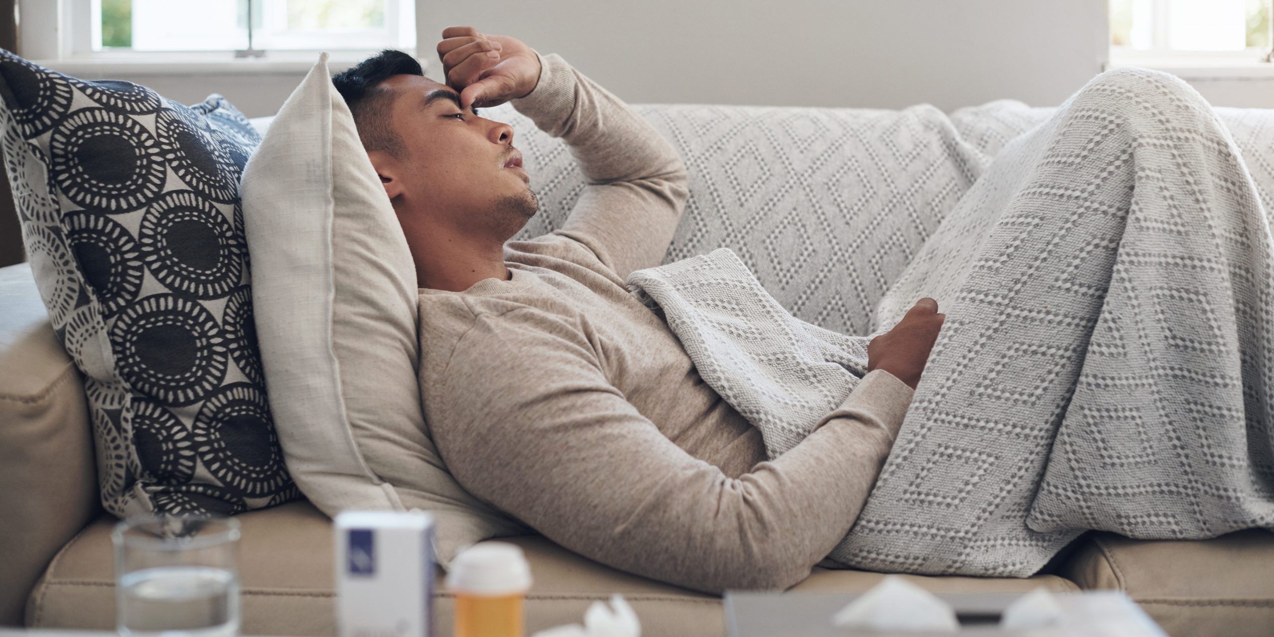 A young man, feeling sick, lays on the couch with water and tissues on a table nearby.