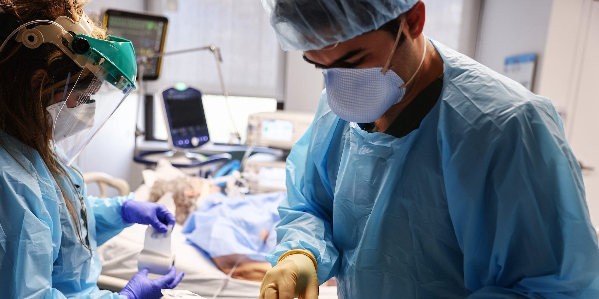 A clinician is looking over his tools while a nursing writes something down. Both are in full PPE. In the background is a COVID-19 patient.