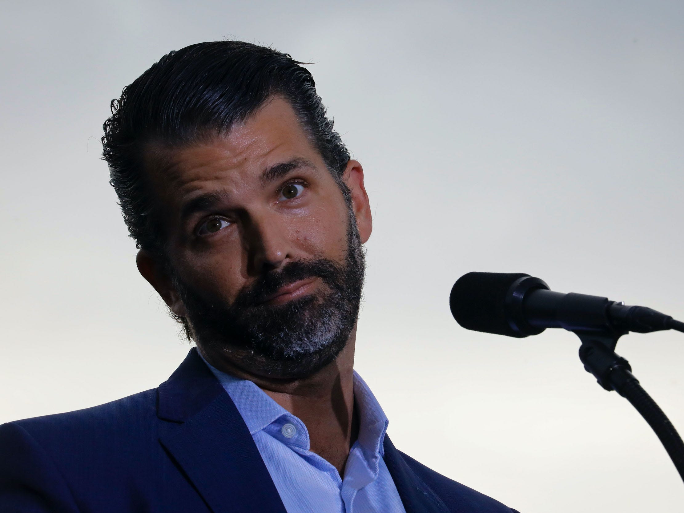 Donald Trump Jr. tilts his head and raises his eyebrows with his lips pursed behind the mic at a Trump rally.