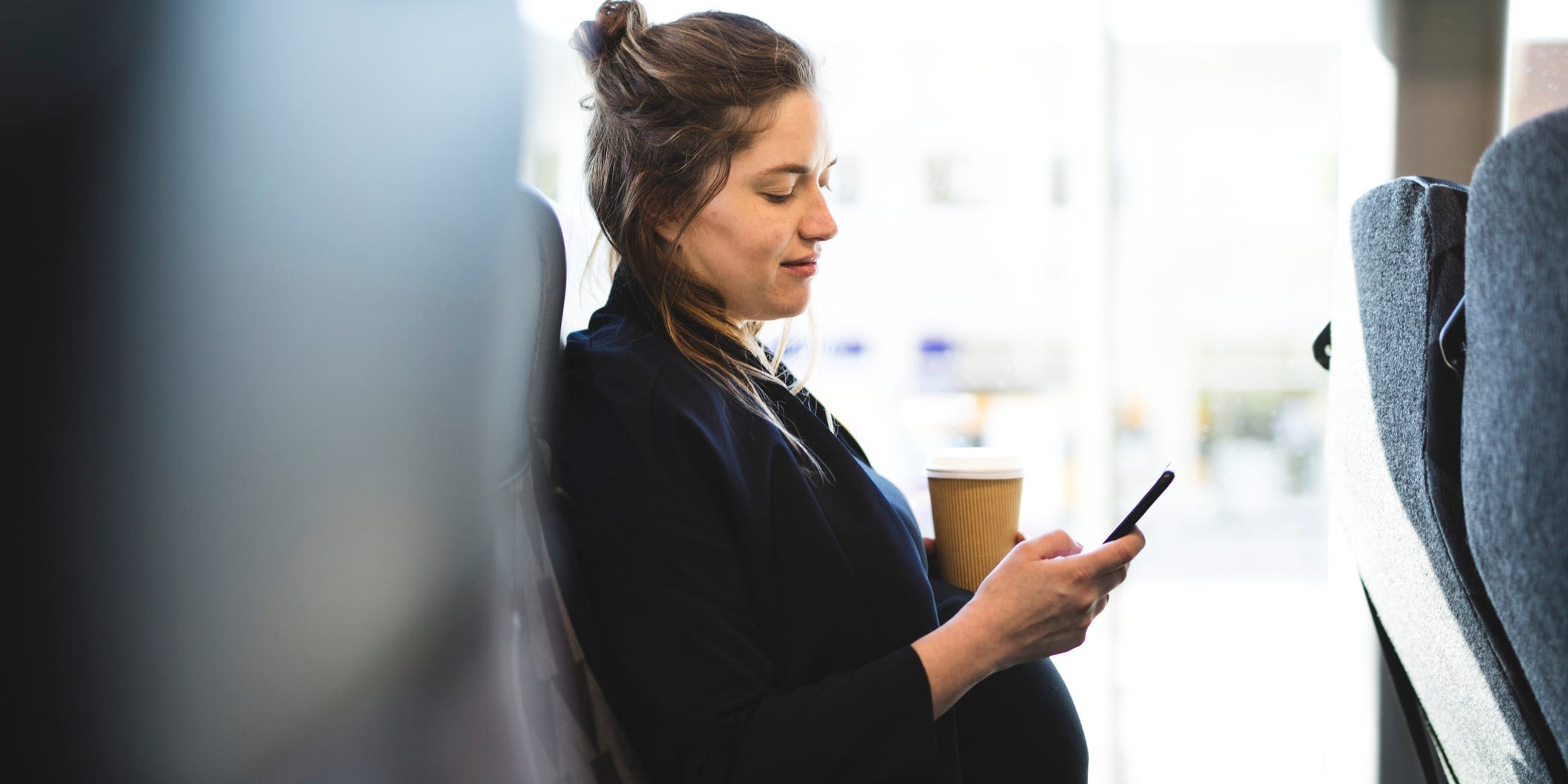 A pregnant woman texts on her phone while holding a cup of coffee.