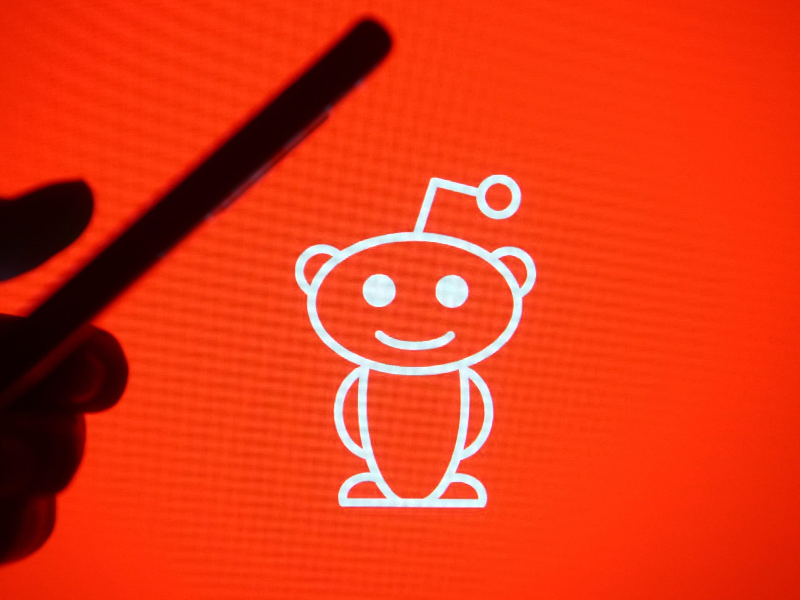 In this photo illustration a silhouette hand holding a mobile phone with a Reddit logo in the background.