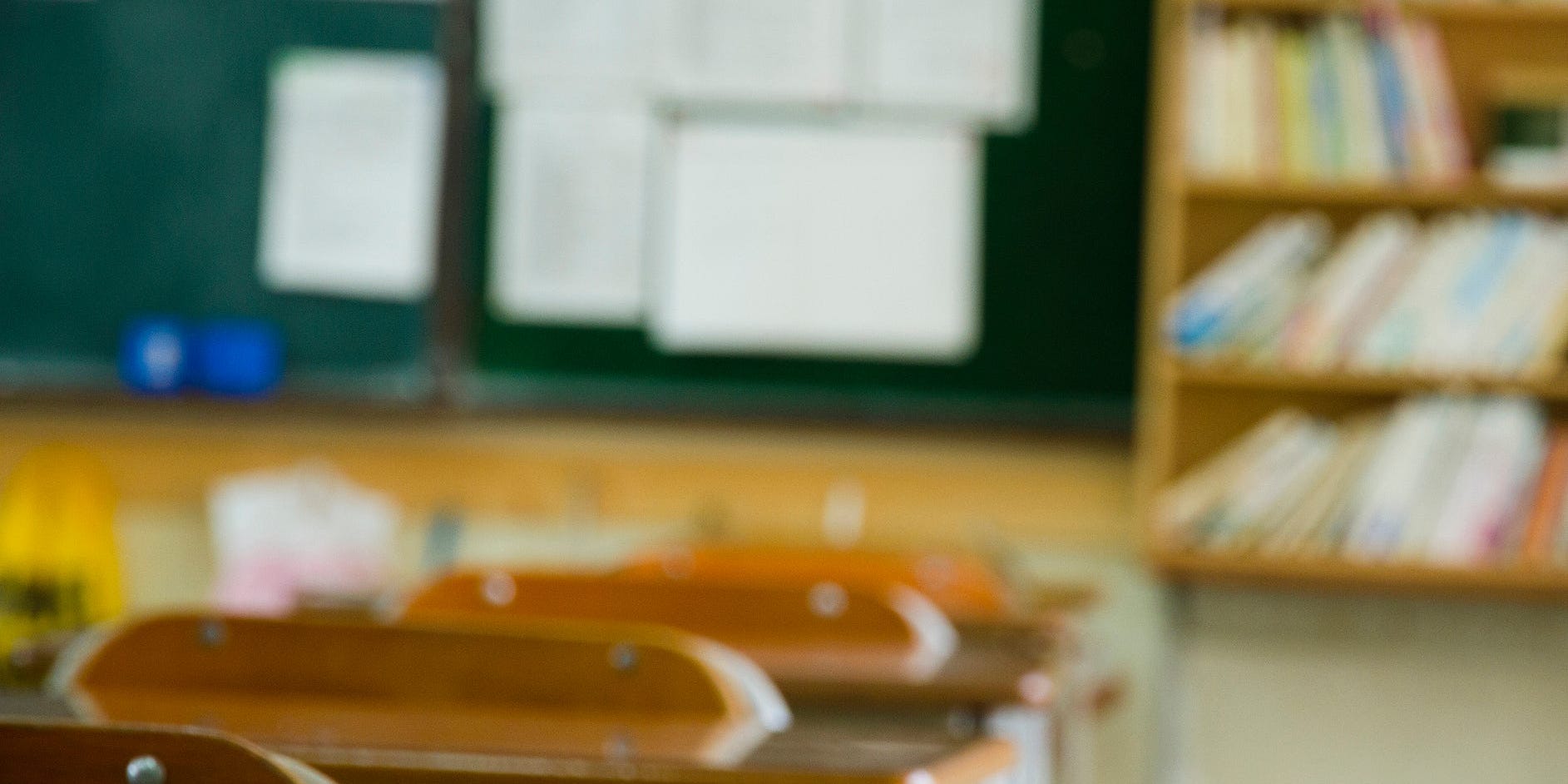 A school classroom, with desks, a blackboard, and bookshelves.