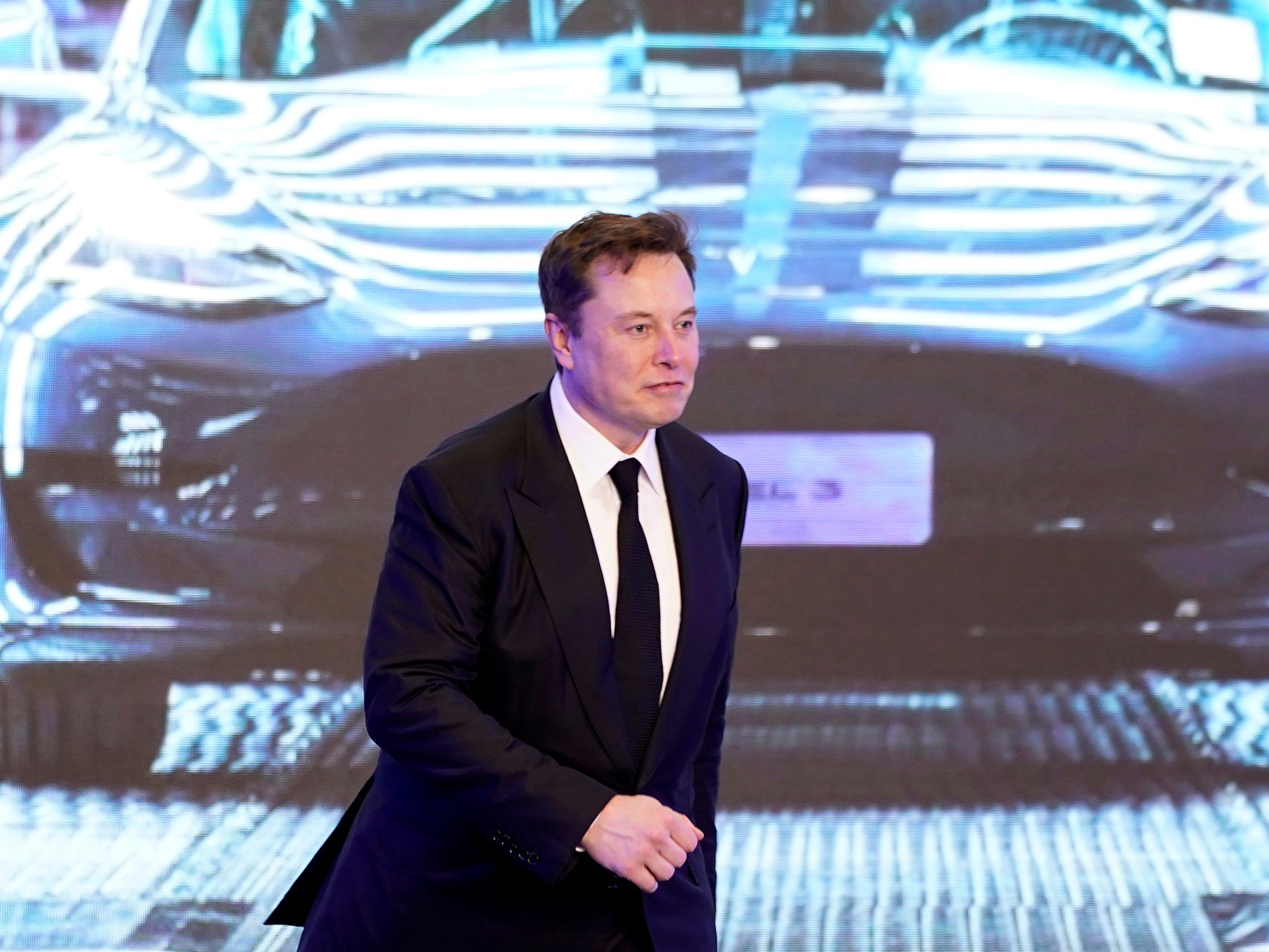 Tesla CEO Elon Musk in a black suit walks on stage in front of an image of a Model Y vehicle