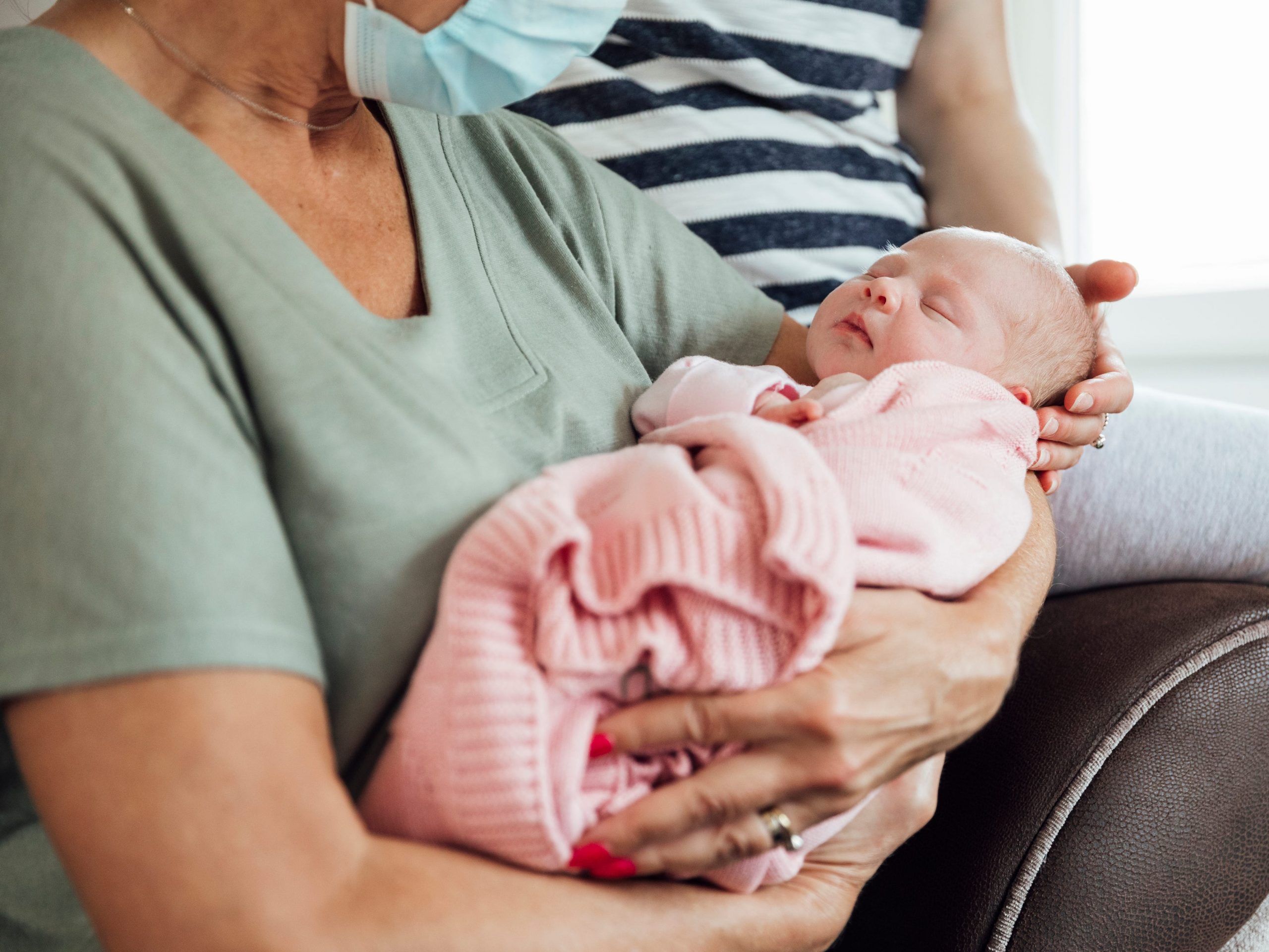 A close-up of a grandmother holding her newborn granddaughter on her lap, with her daughter beside her, the grandmother is wearing a facemask.