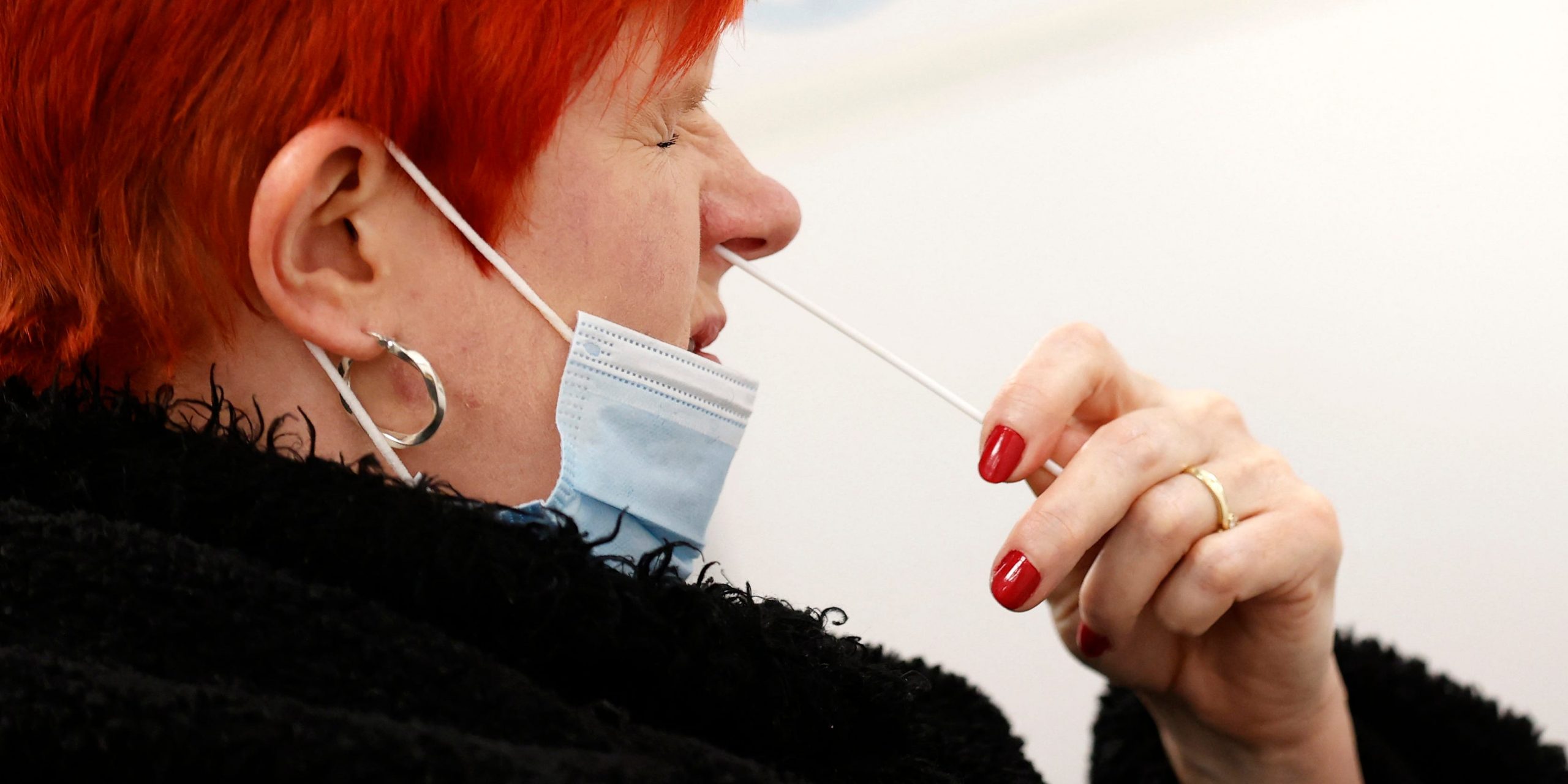 Women with red hair and red nail varnish in a black coat and gold hoops puts COVID-19 test swab into right nostril.