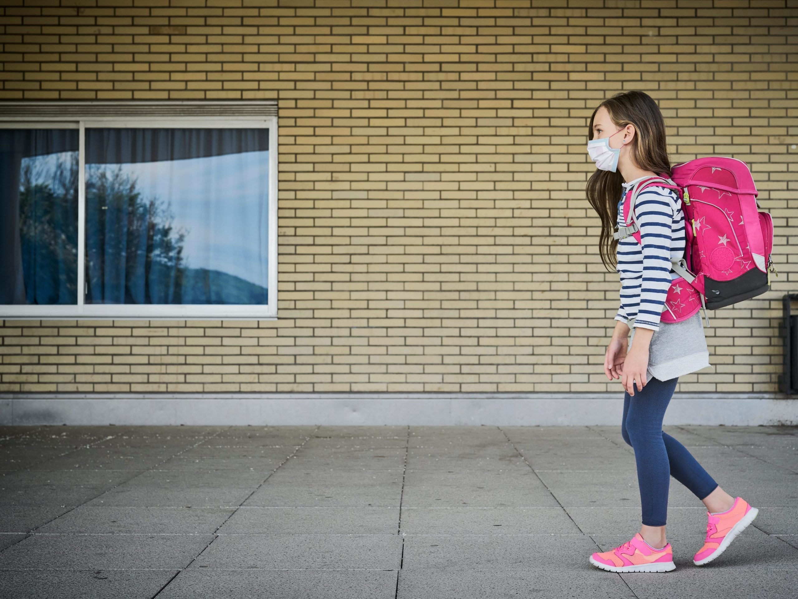 Girl wearing mask and schoolbag walking along building