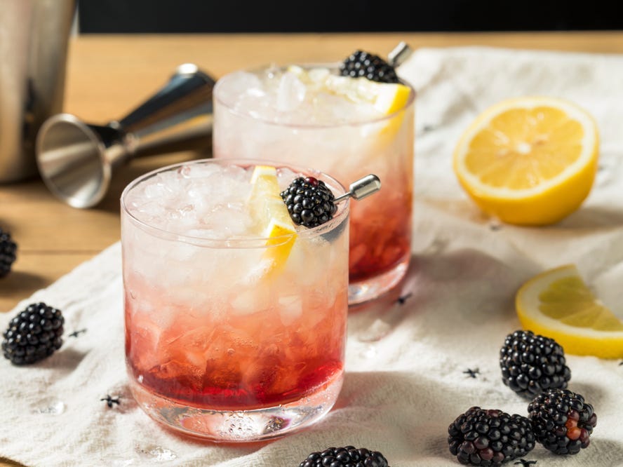 Two glasses of bramble cocktail on a table surrounded by lemons and blackberries.