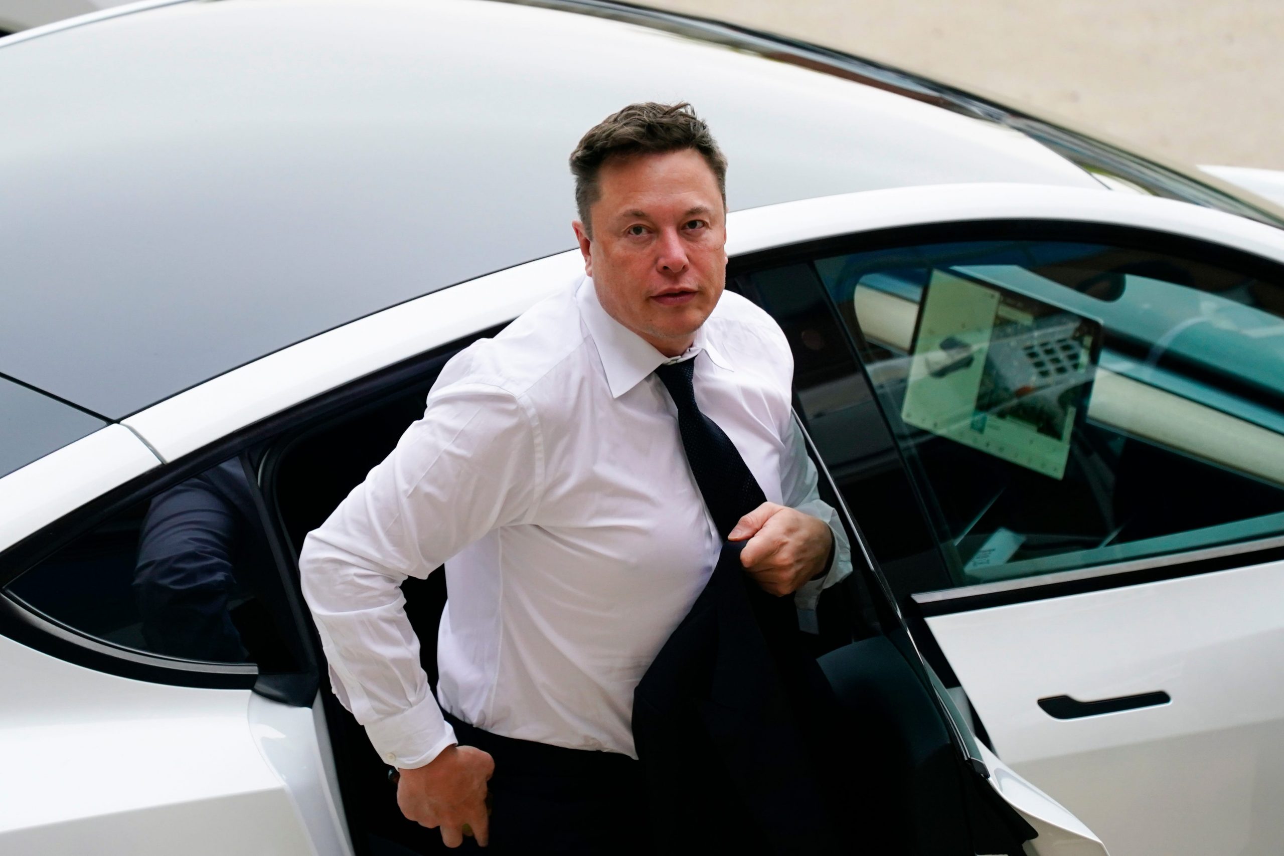 Tesla CEO Elon Musk stepping out of a silver Tesla wearing a white shirt and black tie on a sunny day