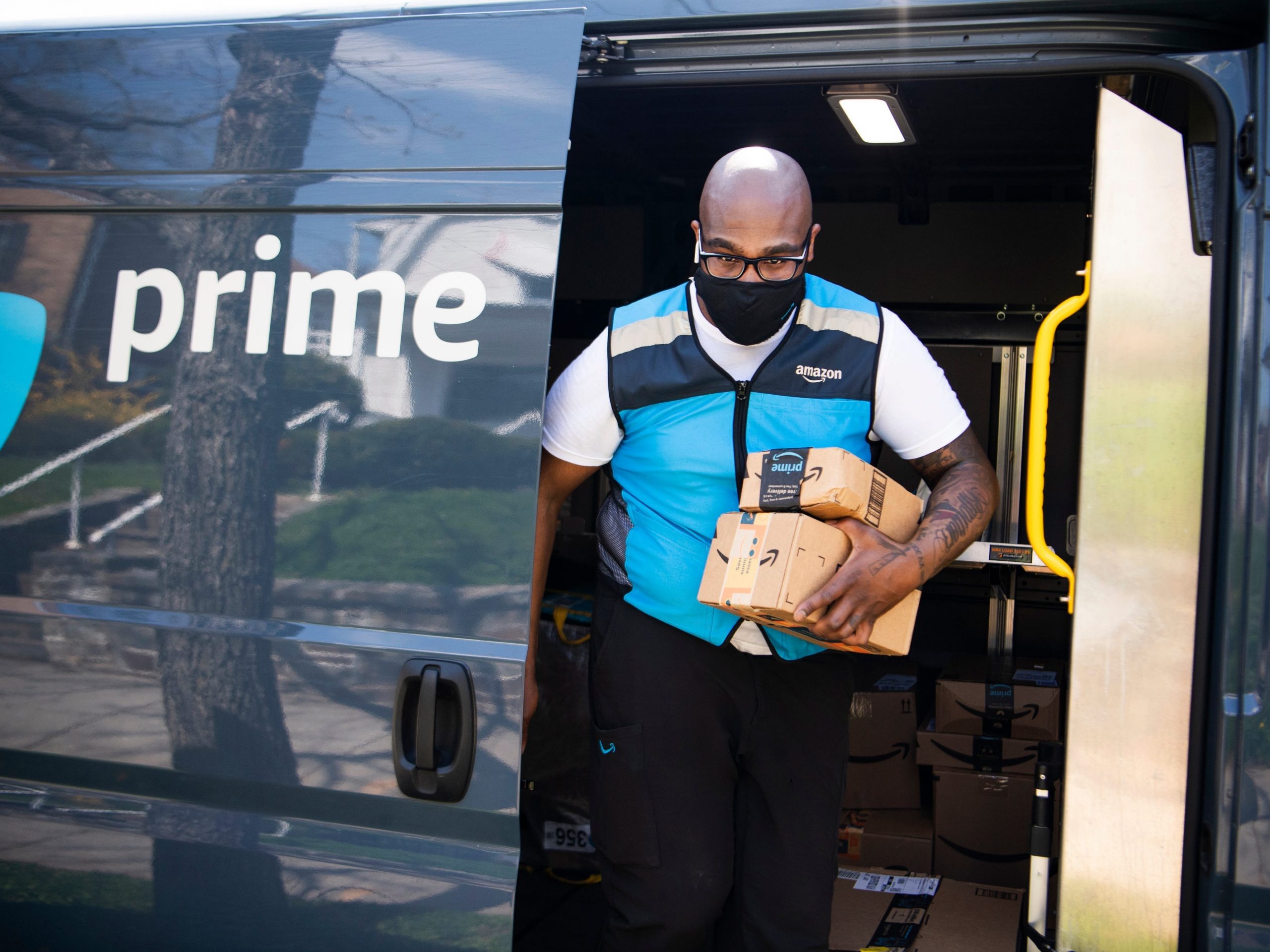 An Amazon prime driver makes a delivery, wearing a blue vest and black face mask and holding two cardboard boxes