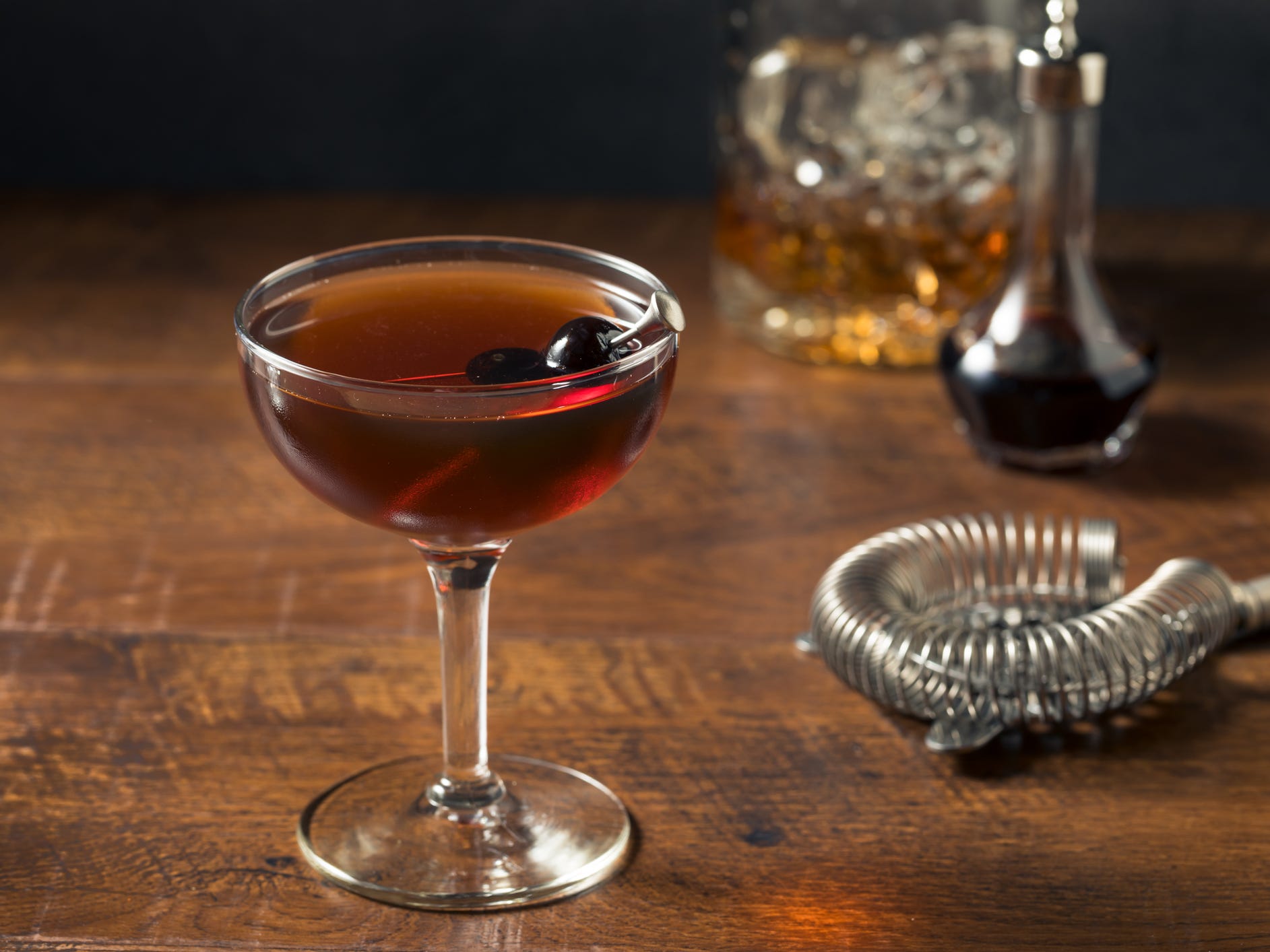 A Manhattan cocktail in a coupe glass garnished with a cherry. A cocktail strainer and a glass decanter of whiskey sit in the background.