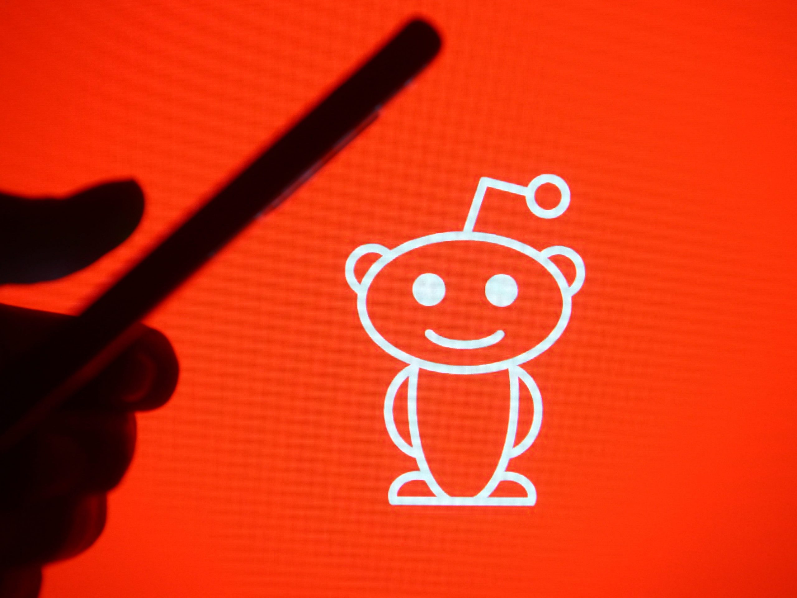 In this photo illustration a silhouette hand holding a mobile phone with a Reddit logo in the background.