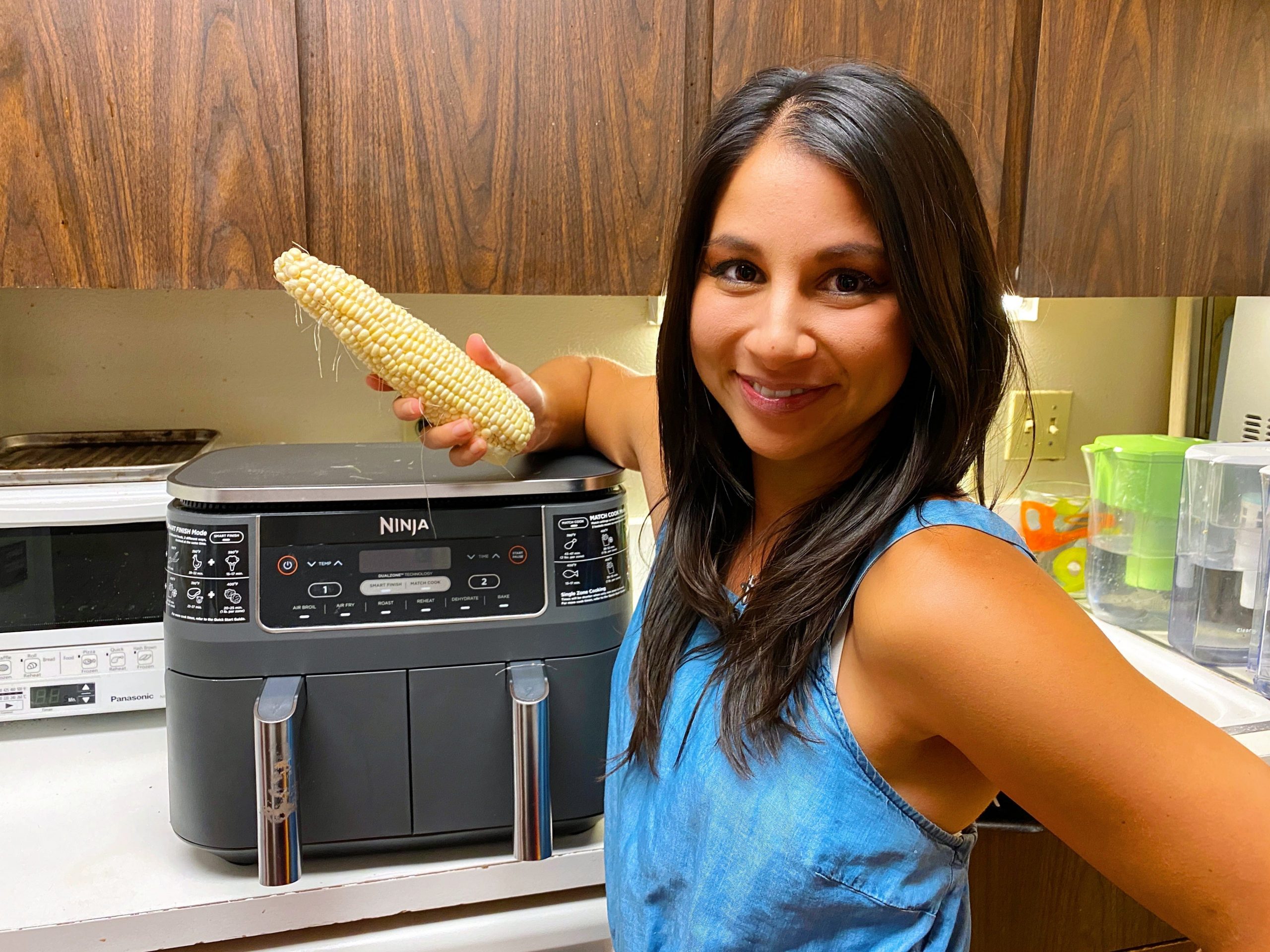 Chelsea holding up a corn on a cob in front of an air fryer.